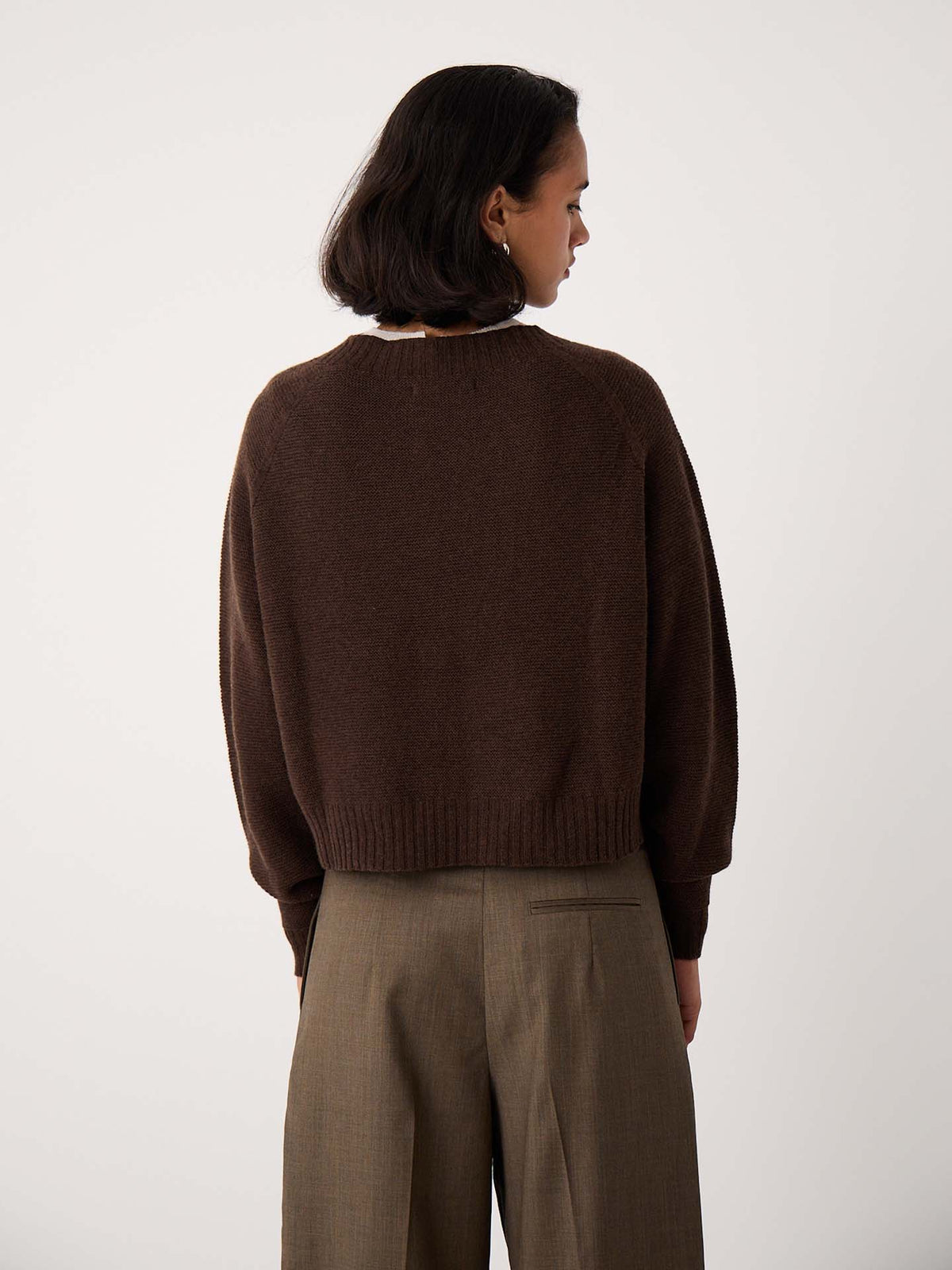 A petite person with short dark hair wears the Poet Knit – Cocoa, a relaxed-fit brown sweater, paired with khaki trousers and stands facing away from the camera against a plain light background.