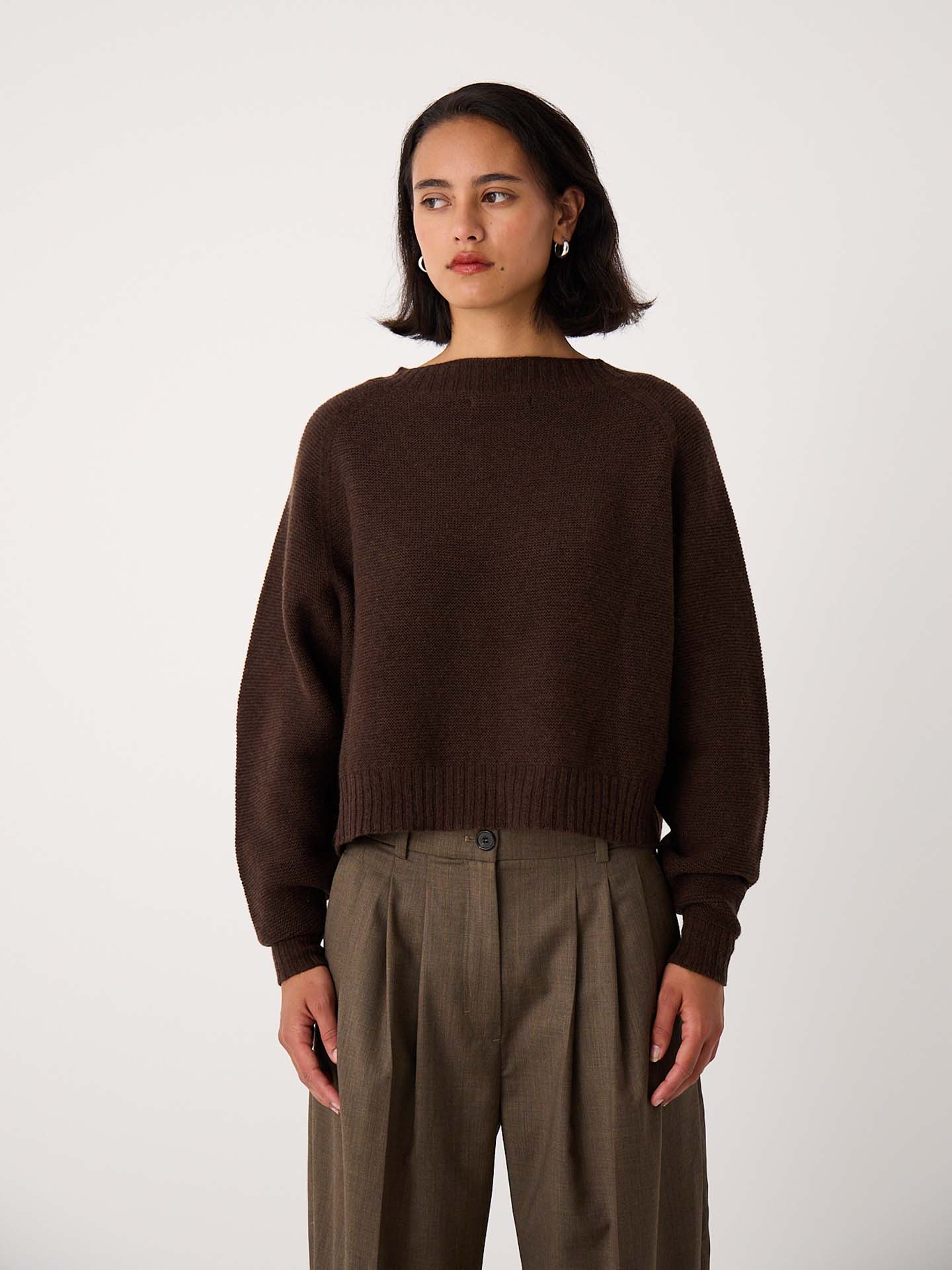 A petite woman with short dark hair wears the Poet Knit in Cocoa, a relaxed fit brown sweater, paired with high-waisted pleated khaki trousers, standing against a plain light background.
