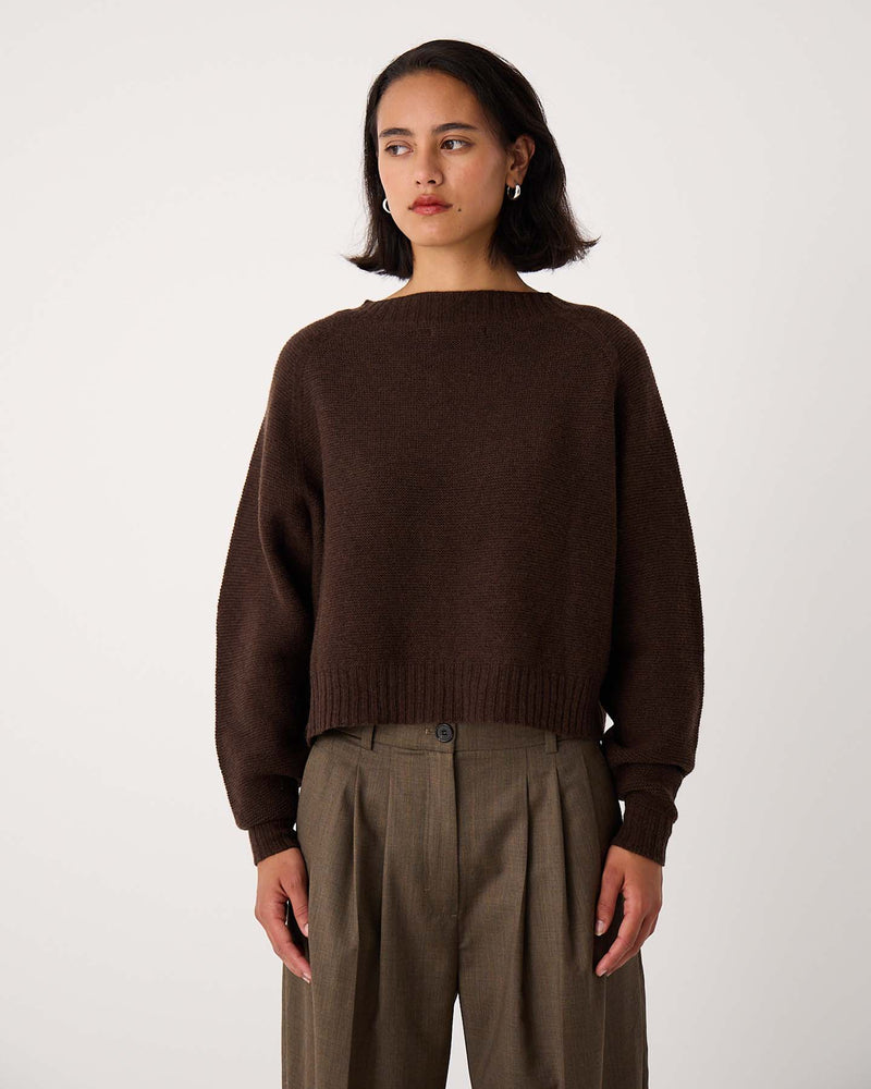A petite woman with short dark hair wears the Poet Knit in Cocoa, a relaxed fit brown sweater, paired with high-waisted pleated khaki trousers, standing against a plain light background.