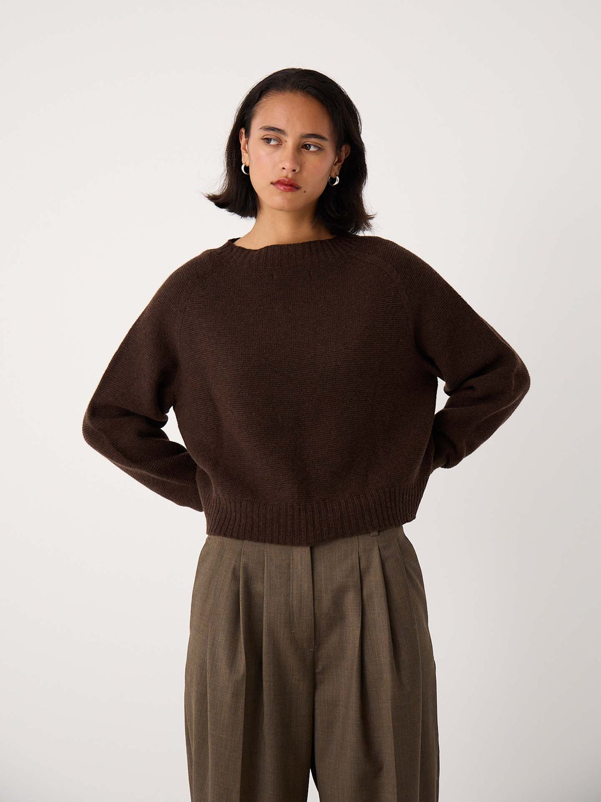 A person wearing the Poet Knit – Cocoa in a cropped style and high-waisted, loose-fitting khaki trousers stands against a plain white background.