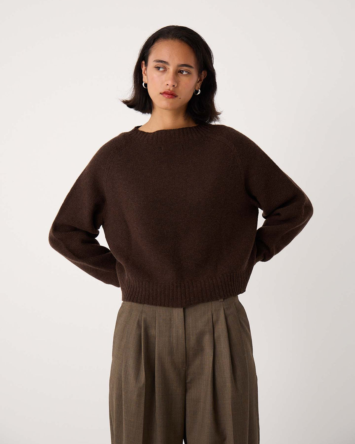 A person wearing the Poet Knit – Cocoa in a cropped style and high-waisted, loose-fitting khaki trousers stands against a plain white background.