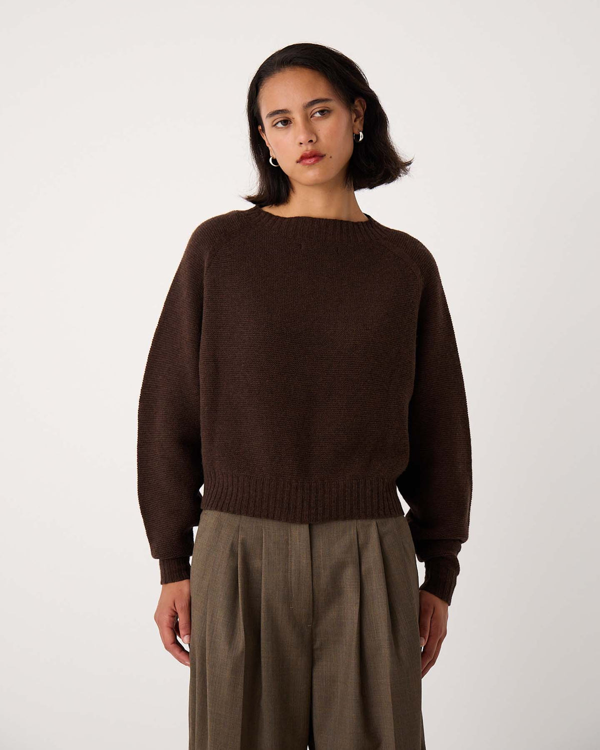 A petite woman with shoulder-length dark hair wears the Poet Knit – Cocoa sweater in a relaxed fit and loose-fitting khaki trousers, standing against a plain white background.