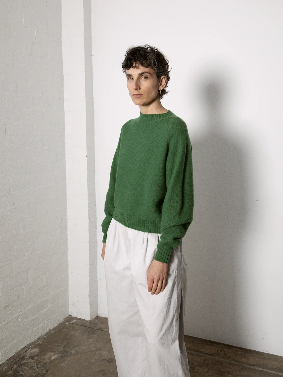 Wearing Francie's Poet Knit in Herb, styled in a relaxed, cropped fit, a person in white pants stands against a white wall, looking towards the camera.