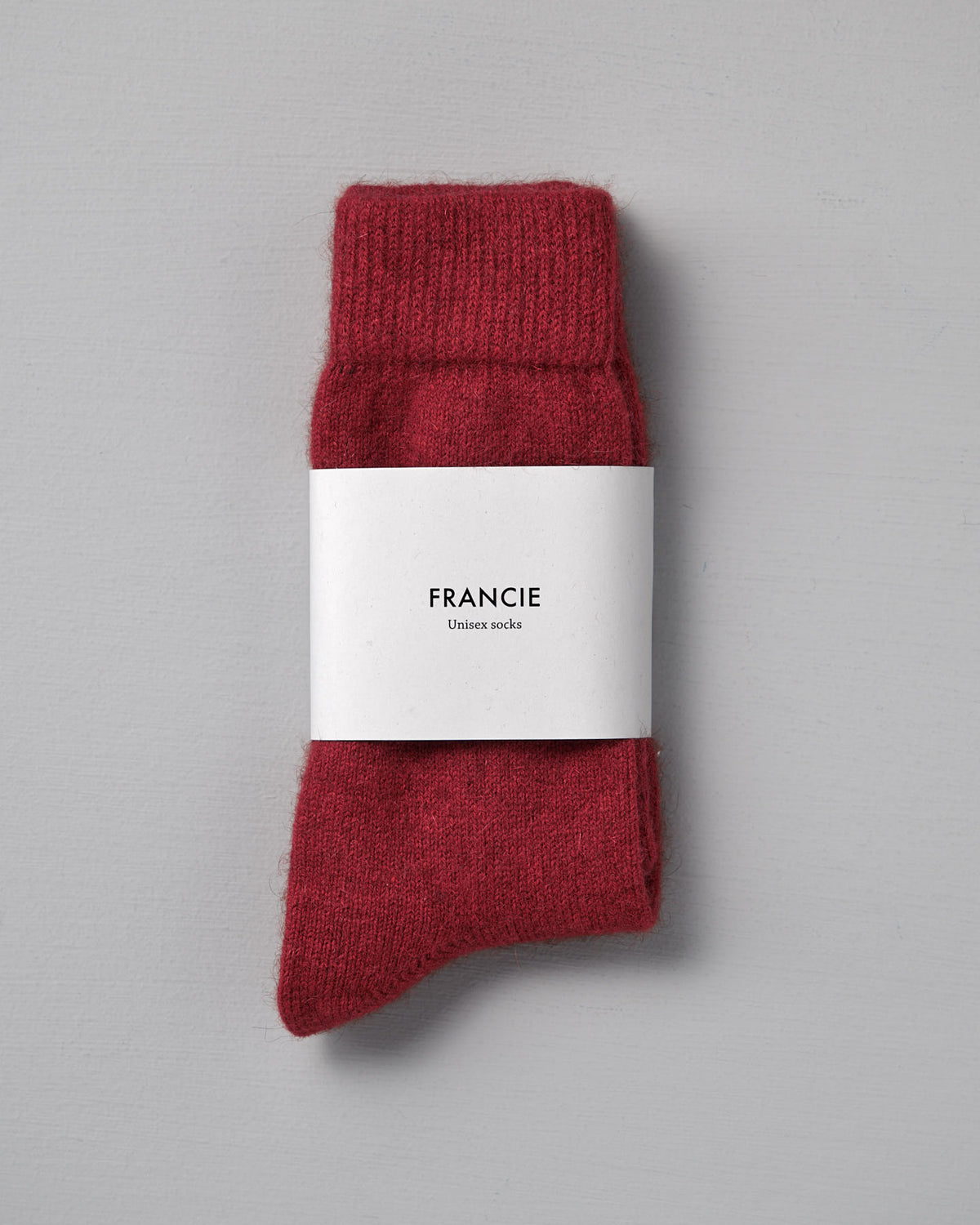 A pair of Francie Possum Merino Socks – Crimson in size Small, folded with a white "Francie Unisex socks" label, placed on a light gray surface.
