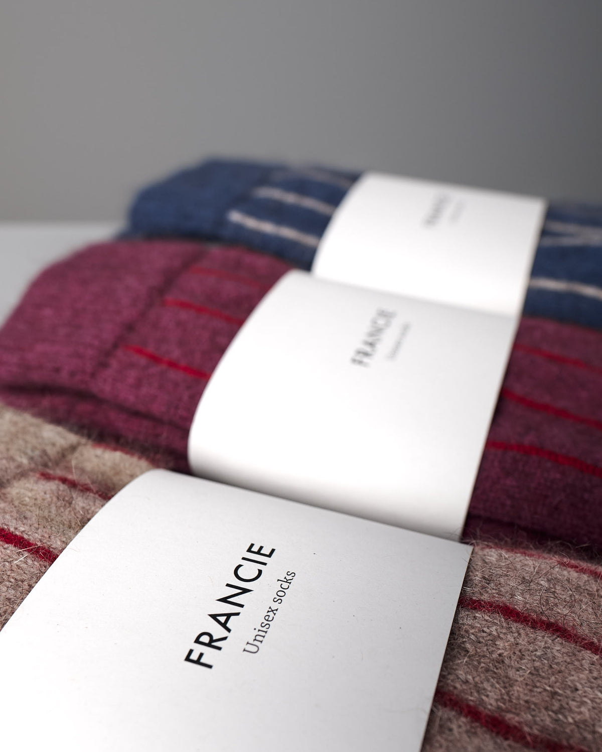 Three pairs of Francie Possum Merino Socks in Natural & Poppy Stripe are stacked, each with a white brand label. Refer to our size chart for EU, UK, and US sizes available in small, medium, or large.