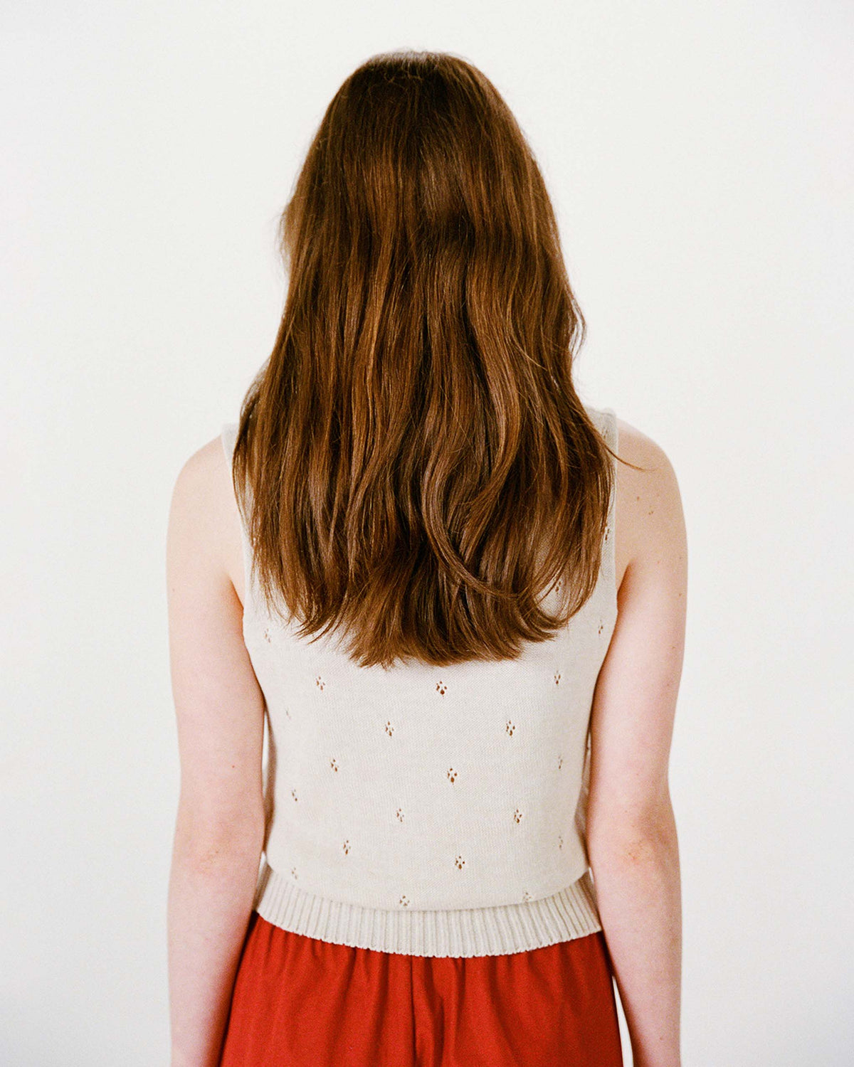 A person with long brown hair, wearing a sleeveless beige top and a red skirt, stands facing away against a plain white background.