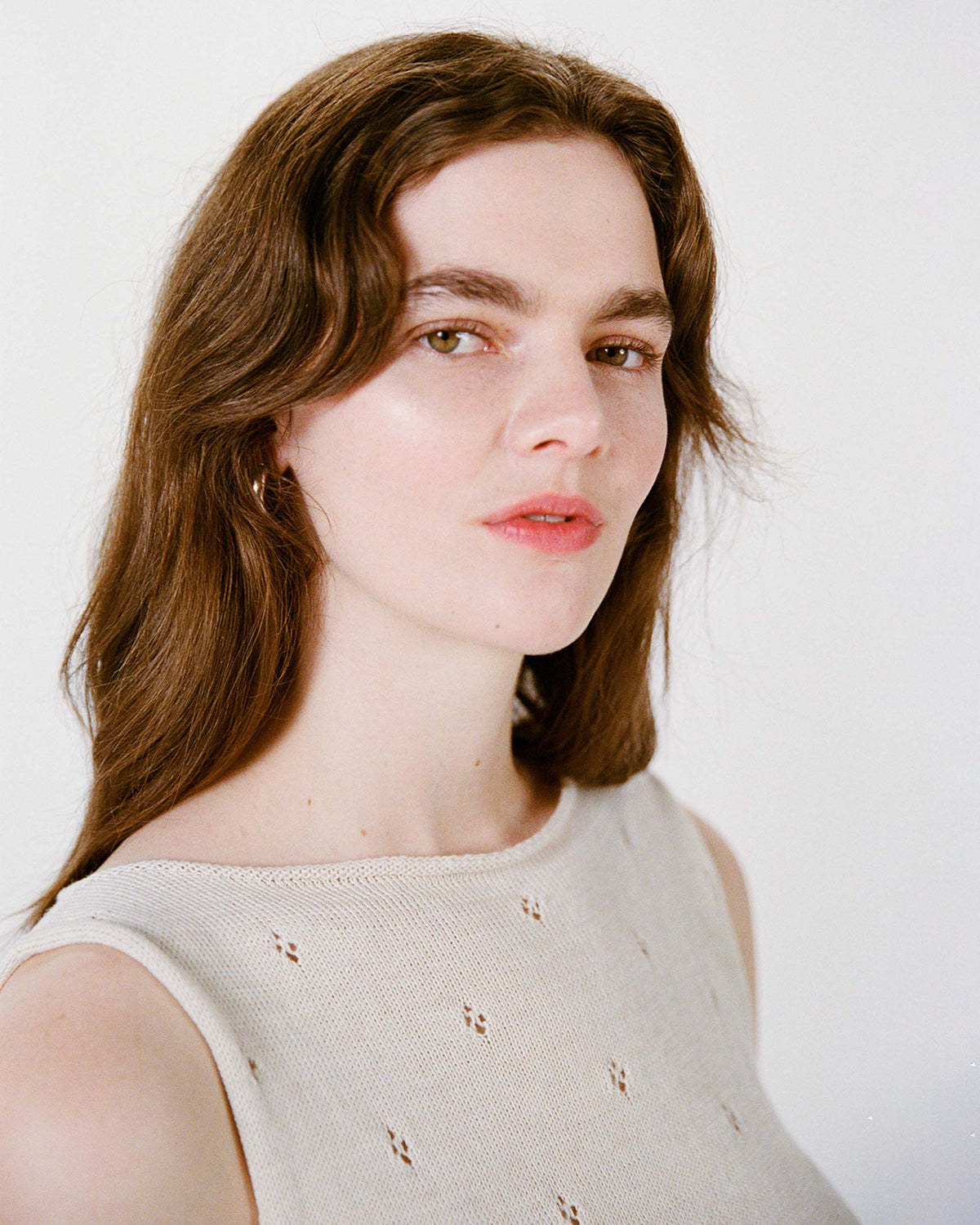 A woman with long brown hair and light skin wears a sleeveless beige top and looks slightly to the side against a plain white background.