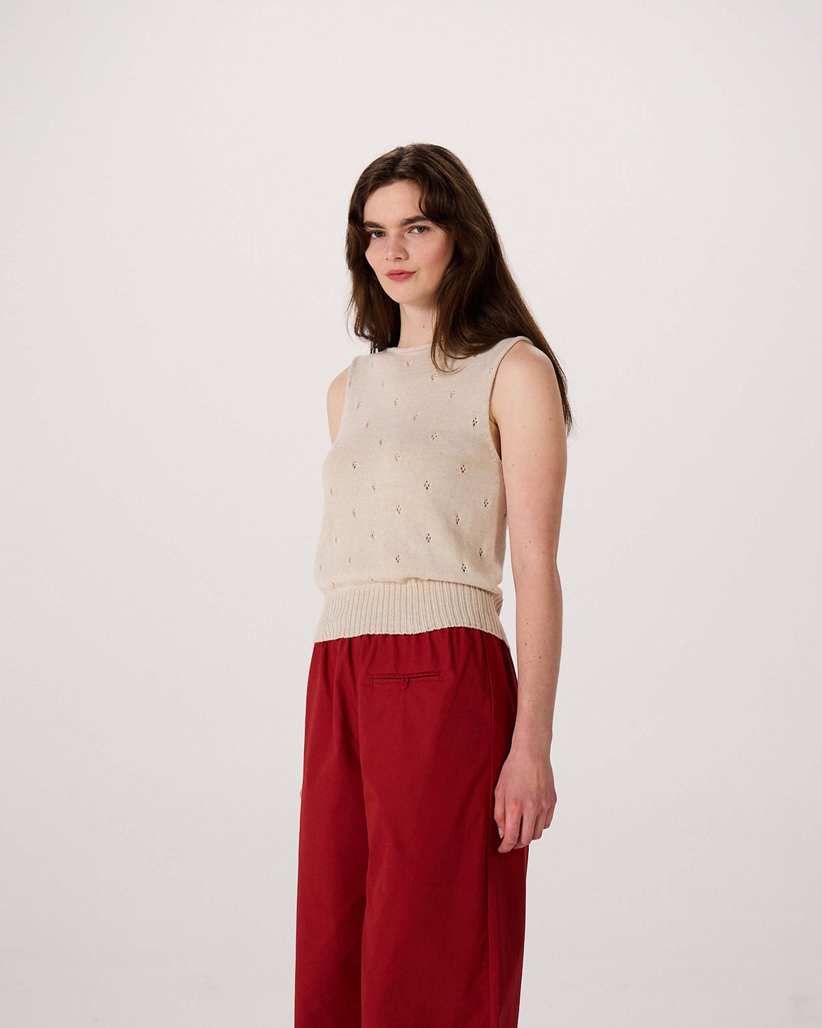 A woman with long brown hair wears a sleeveless beige sweater and red pants, standing against a plain white background.