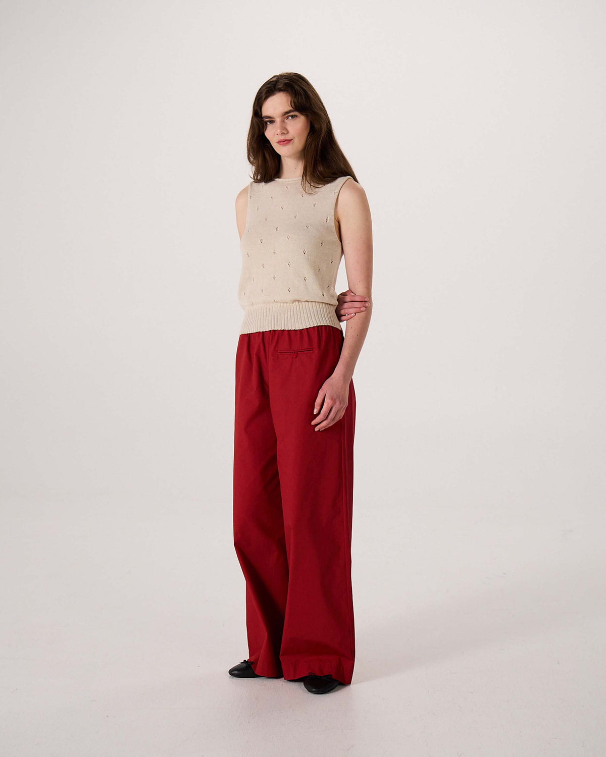 A person stands against a plain background, wearing a sleeveless beige top, loose red pants, and black shoes.