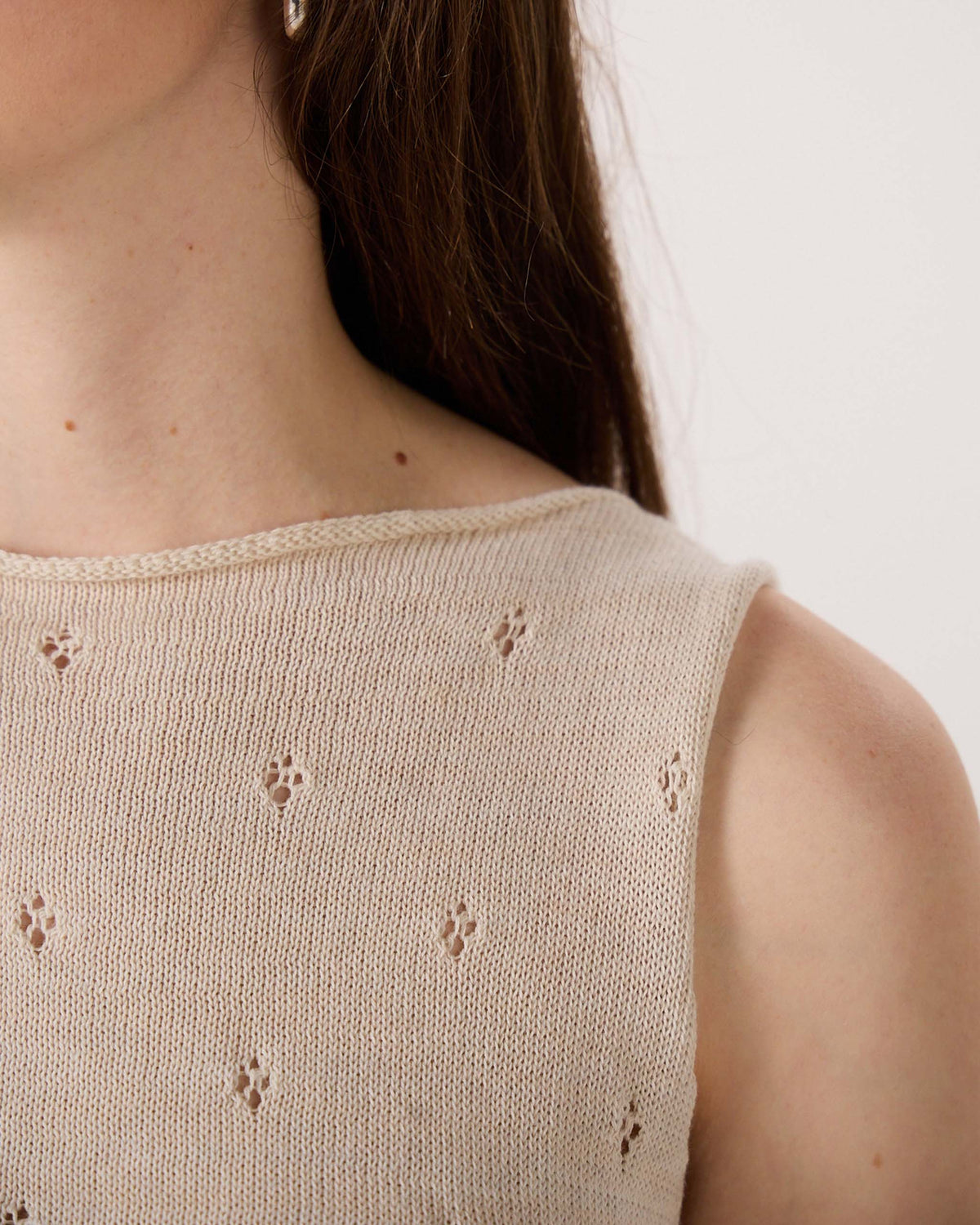 Close-up of a person wearing a sleeveless beige knit top with small decorative holes; part of the face and shoulder are visible against a plain background.