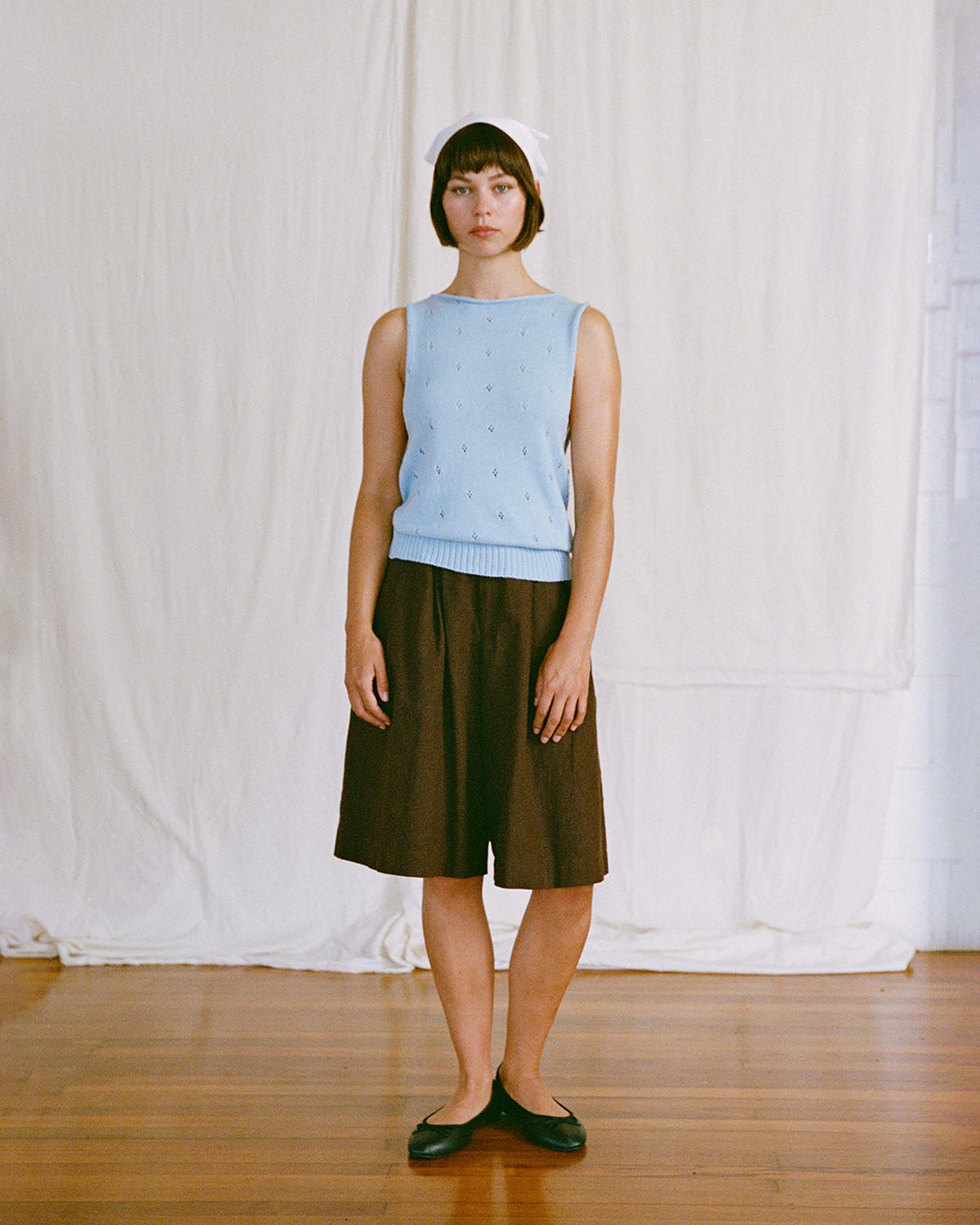 A woman stands indoors on a wooden floor, wearing a light blue sleeveless top, brown knee-length shorts, black flats, and a white headscarf. A white sheet hangs in the background.