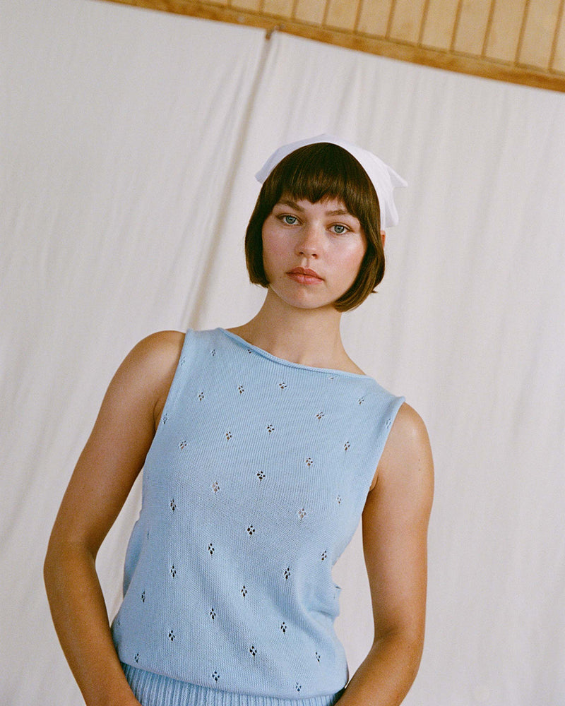 Woman with short dark hair and a white headscarf wears a sleeveless light blue top, standing indoors against a neutral background.