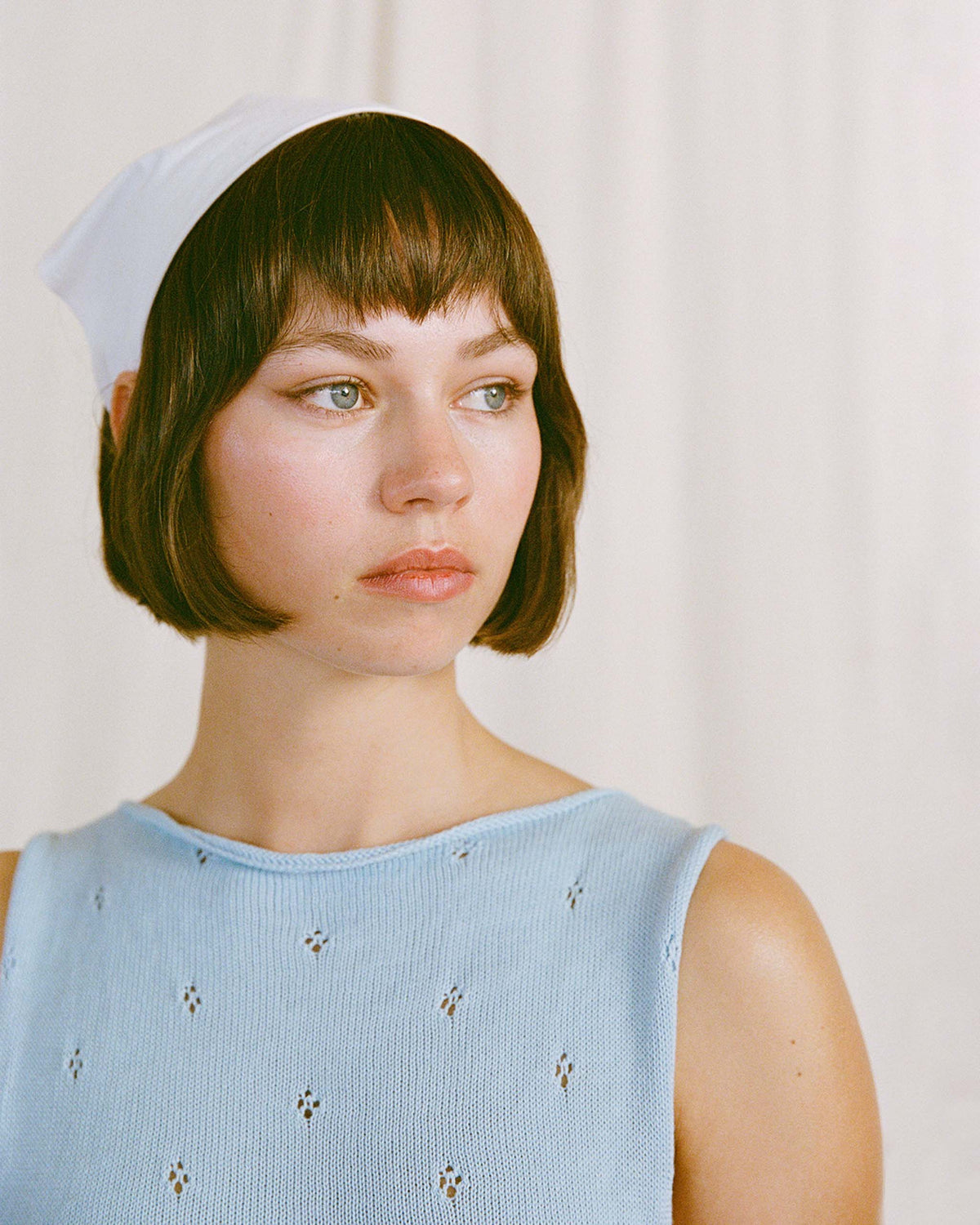 A woman with short brown hair, wearing a light blue sleeveless top and a white headscarf, looks to the side in front of a plain light background.