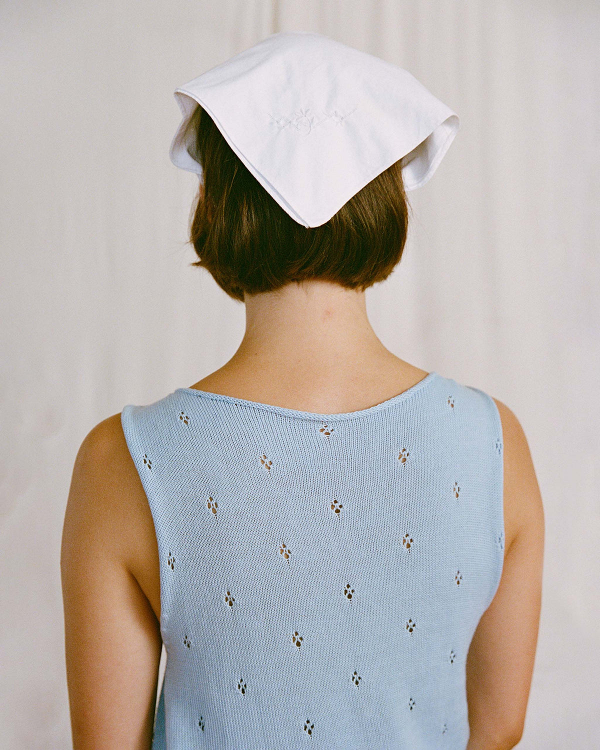 A person with short brown hair, seen from behind, wears a light blue sleeveless top with small holes and a white cloth or kerchief covering their head.