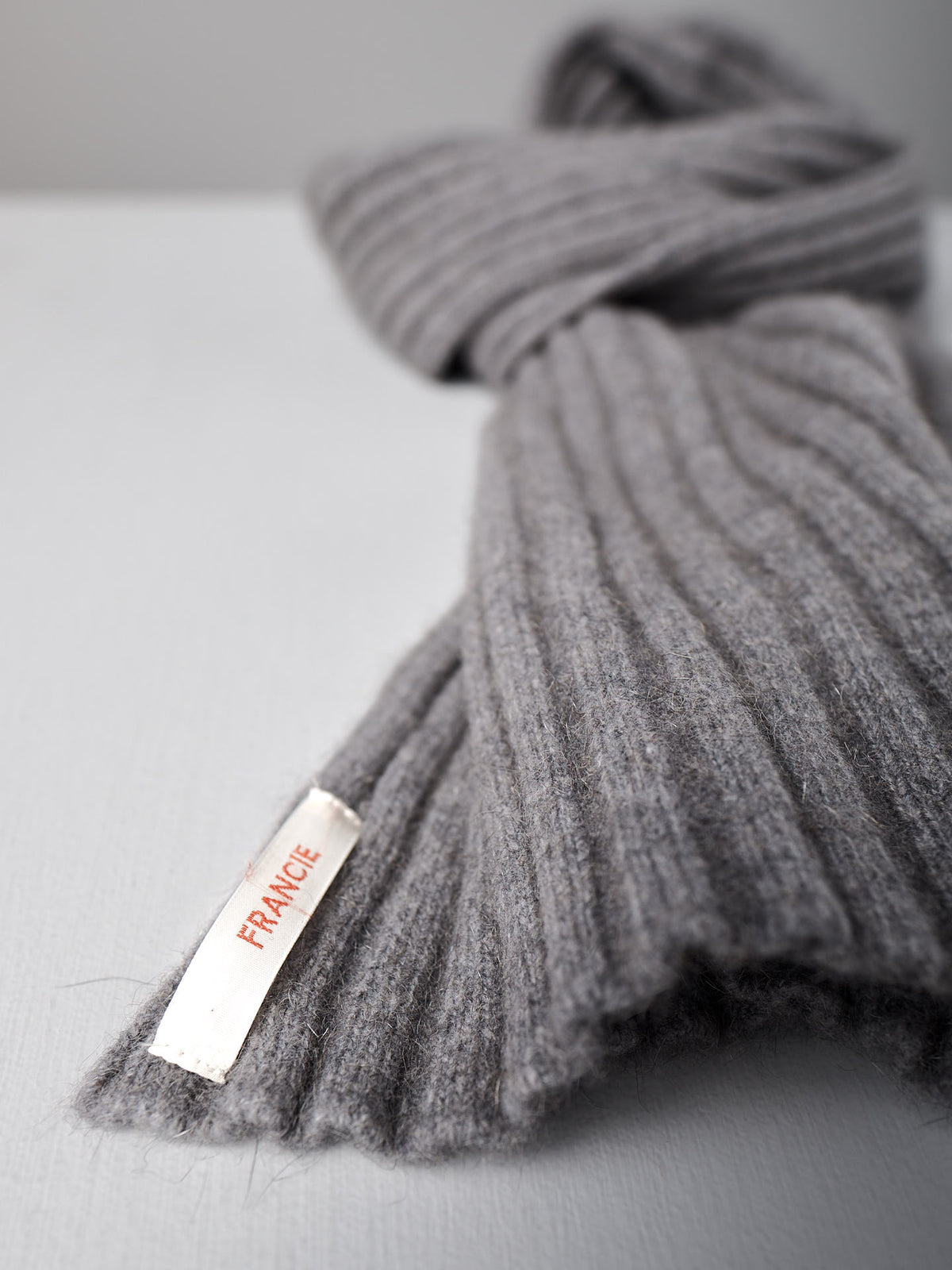 A ribbed herb green wool Ripple Scarf by Francie, 24cm wide and 170cm long, with a white label reading &quot;FRANCE&quot; in red, lies on a light grey surface.