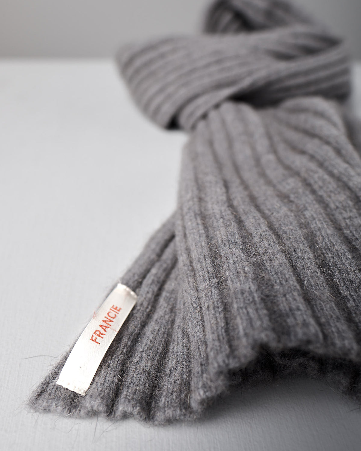 A ribbed herb green wool Ripple Scarf by Francie, 24cm wide and 170cm long, with a white label reading "FRANCE" in red, lies on a light grey surface.