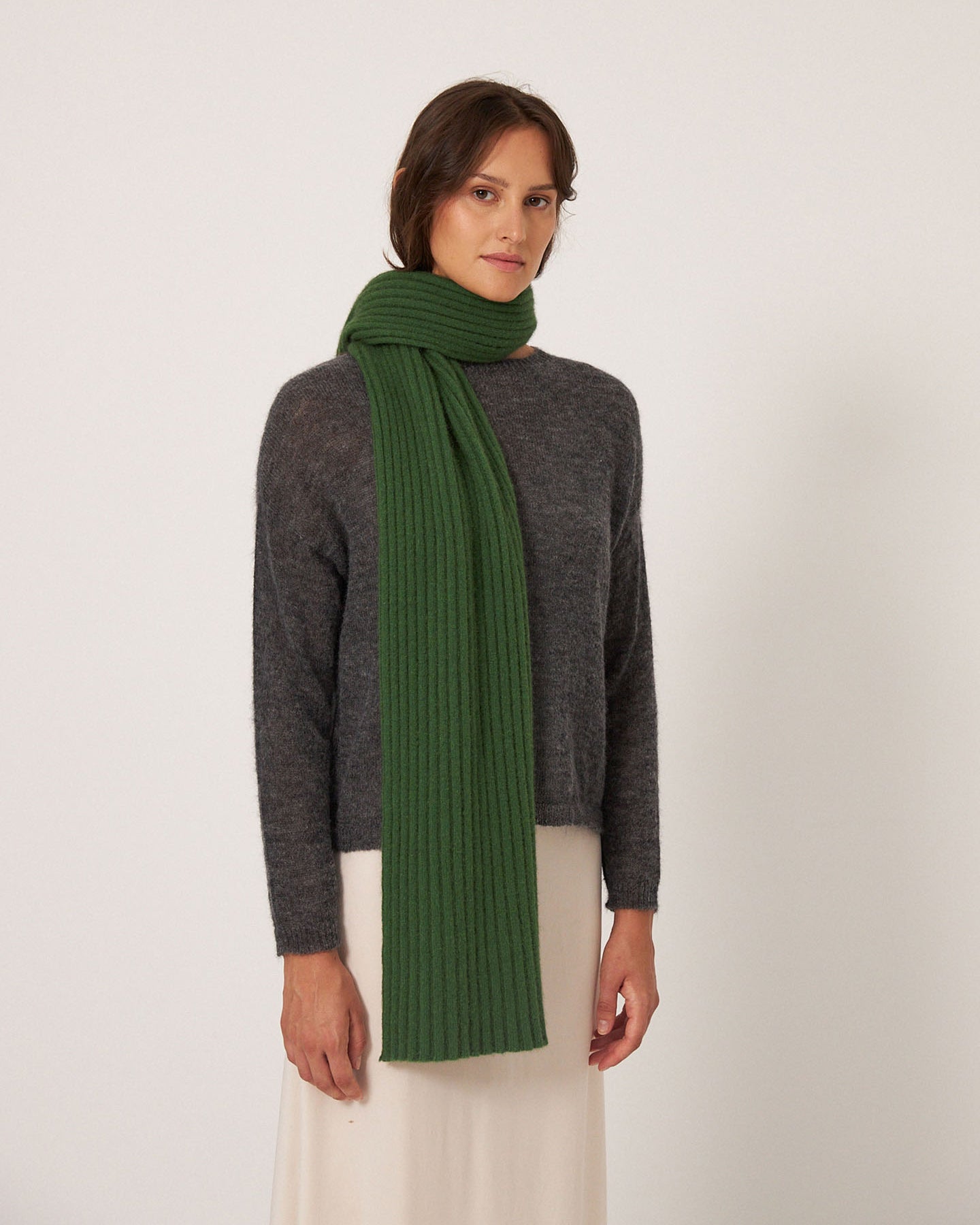 A person wearing Francie's Ripple Scarf – Herb Green (170cm x 24cm), a dark gray sweater, and a light skirt stands against a plain white background.