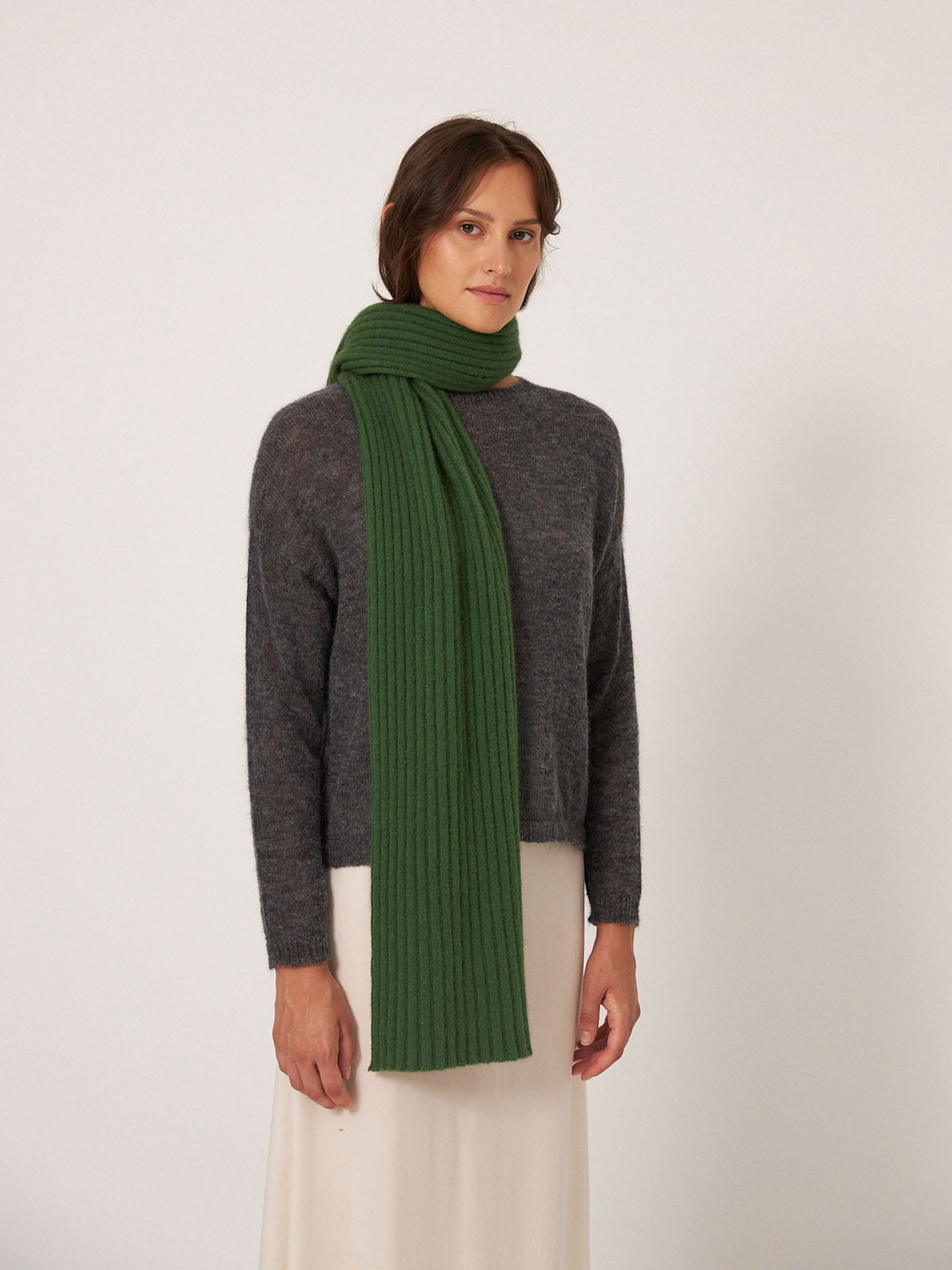 A person wearing Francie's Ripple Scarf – Herb Green (170cm x 24cm), a dark gray sweater, and a light skirt stands against a plain white background.