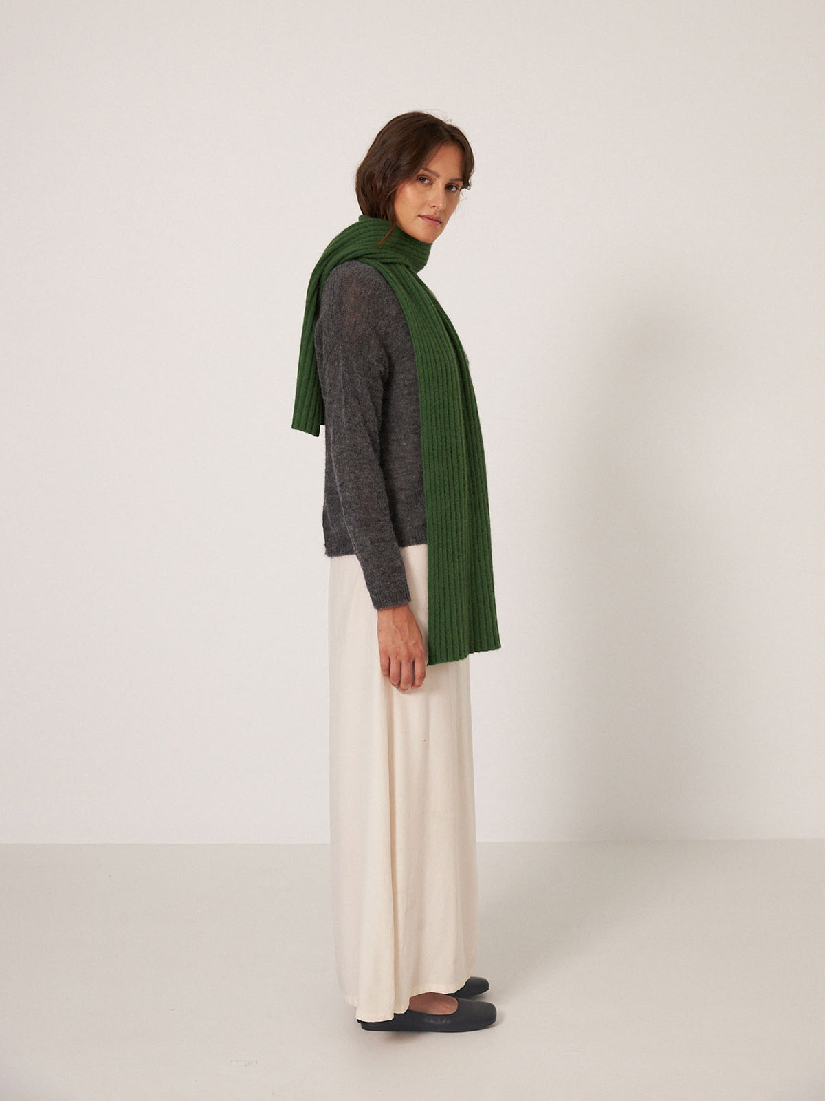 A person stands sideways against a plain white background wearing Francie’s Ripple Scarf in Herb Green, a long grey sweater, a white skirt, and black shoes. The scarf measures 170cm long by 24cm wide.