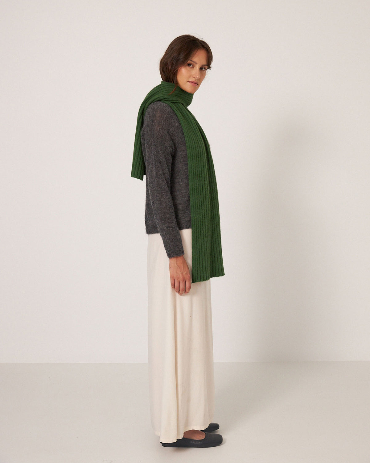 A person stands sideways against a plain white background wearing Francie’s Ripple Scarf in Herb Green, a long grey sweater, a white skirt, and black shoes. The scarf measures 170cm long by 24cm wide.