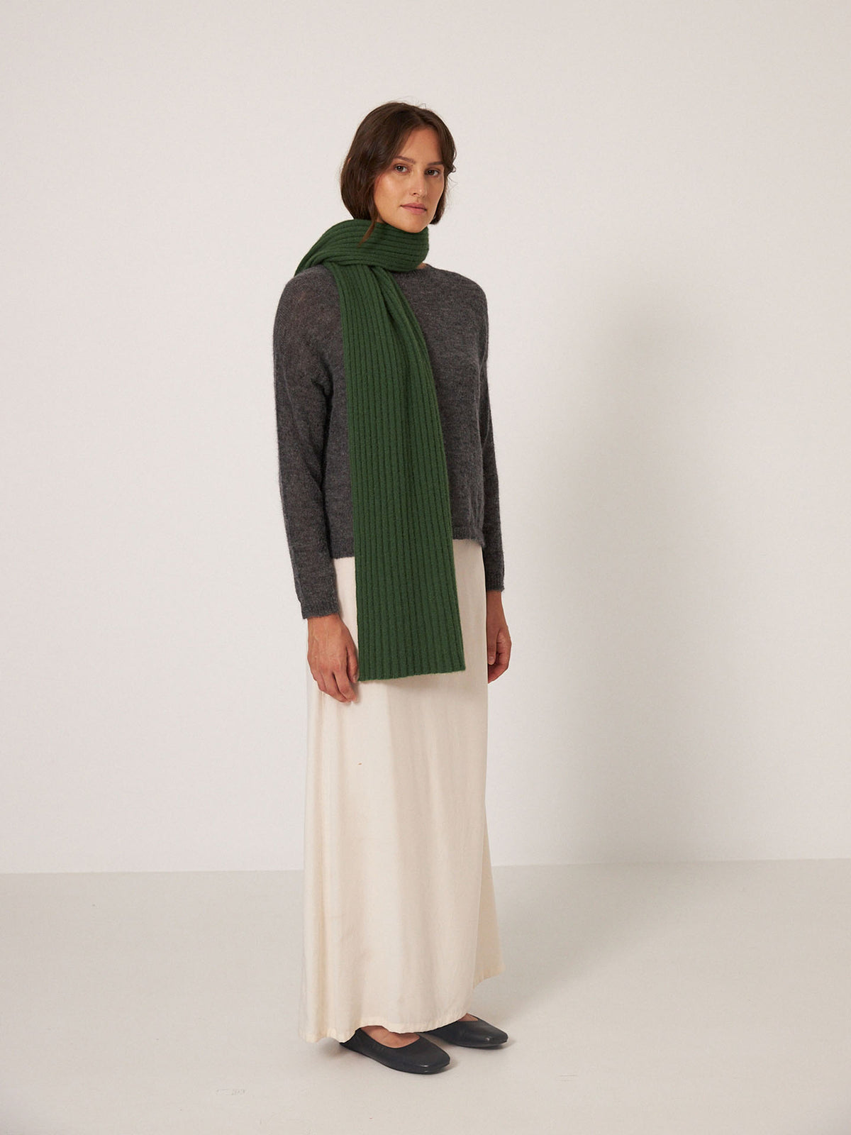 A person stands against a plain background wearing a grey sweater, Francie&#39;s Ripple Scarf in Herb Green (24cm x 170cm), a cream-colored skirt, and black shoes.