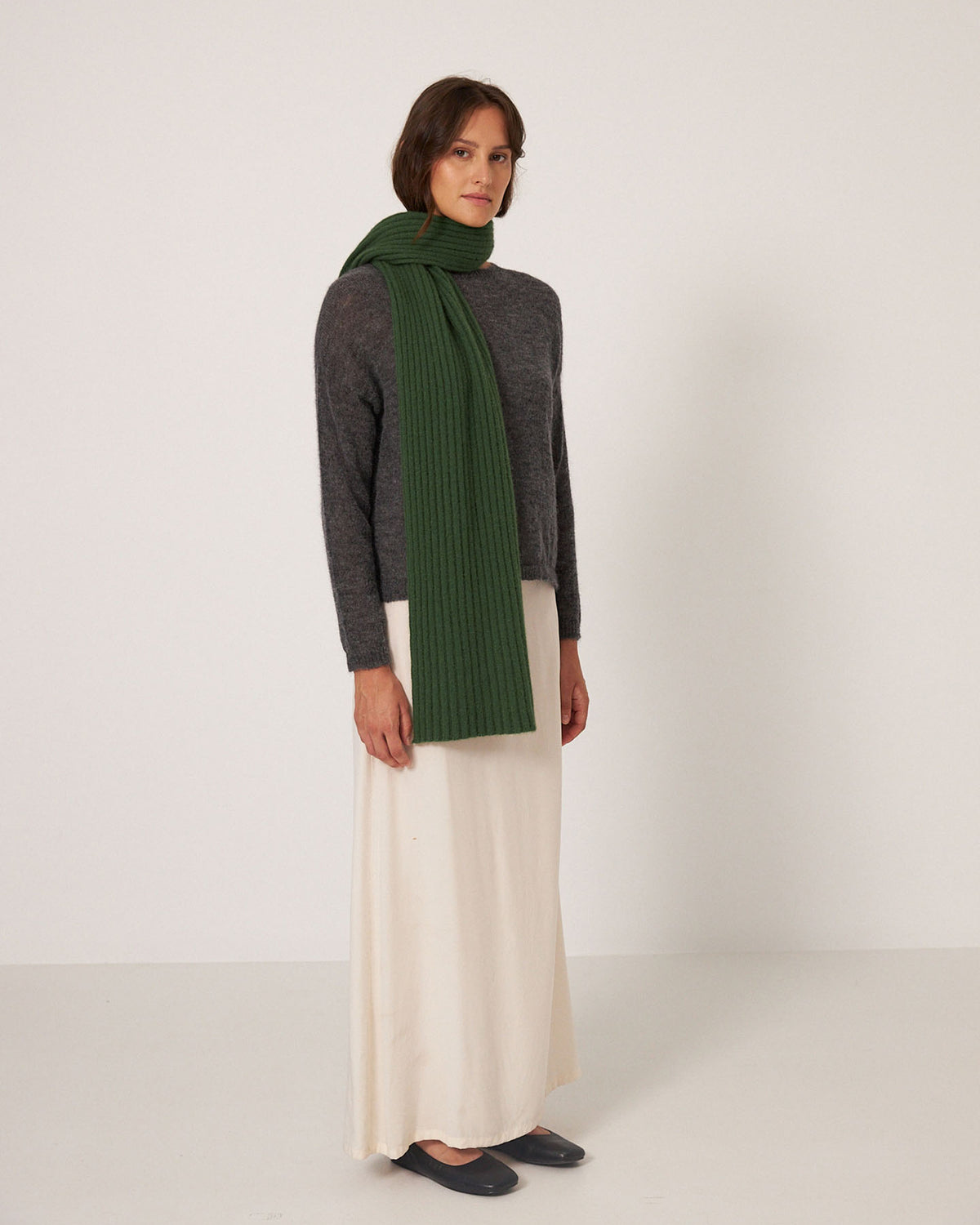 A person stands against a plain background wearing a grey sweater, Francie's Ripple Scarf in Herb Green (24cm x 170cm), a cream-colored skirt, and black shoes.