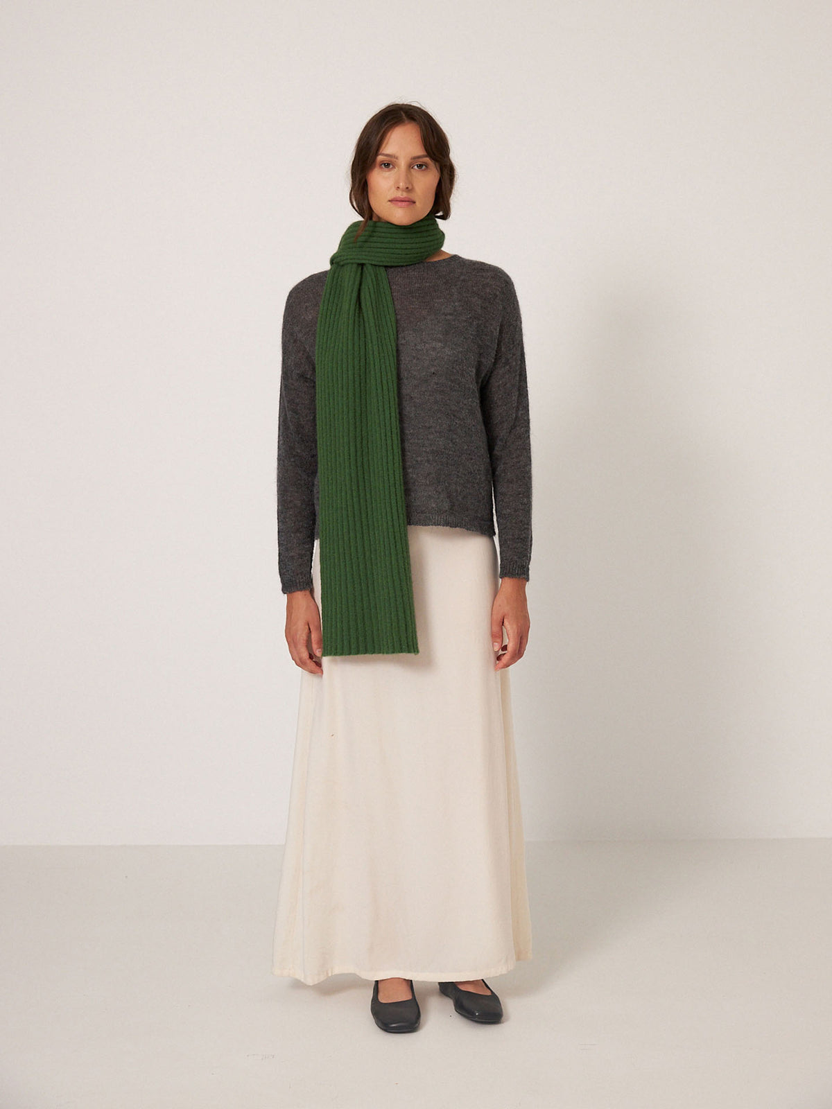 A person stands facing forward against a plain, light background, wearing a gray sweater, long cream skirt, black shoes, and the Francie Ripple Scarf – Herb Green (170cm at widest point).