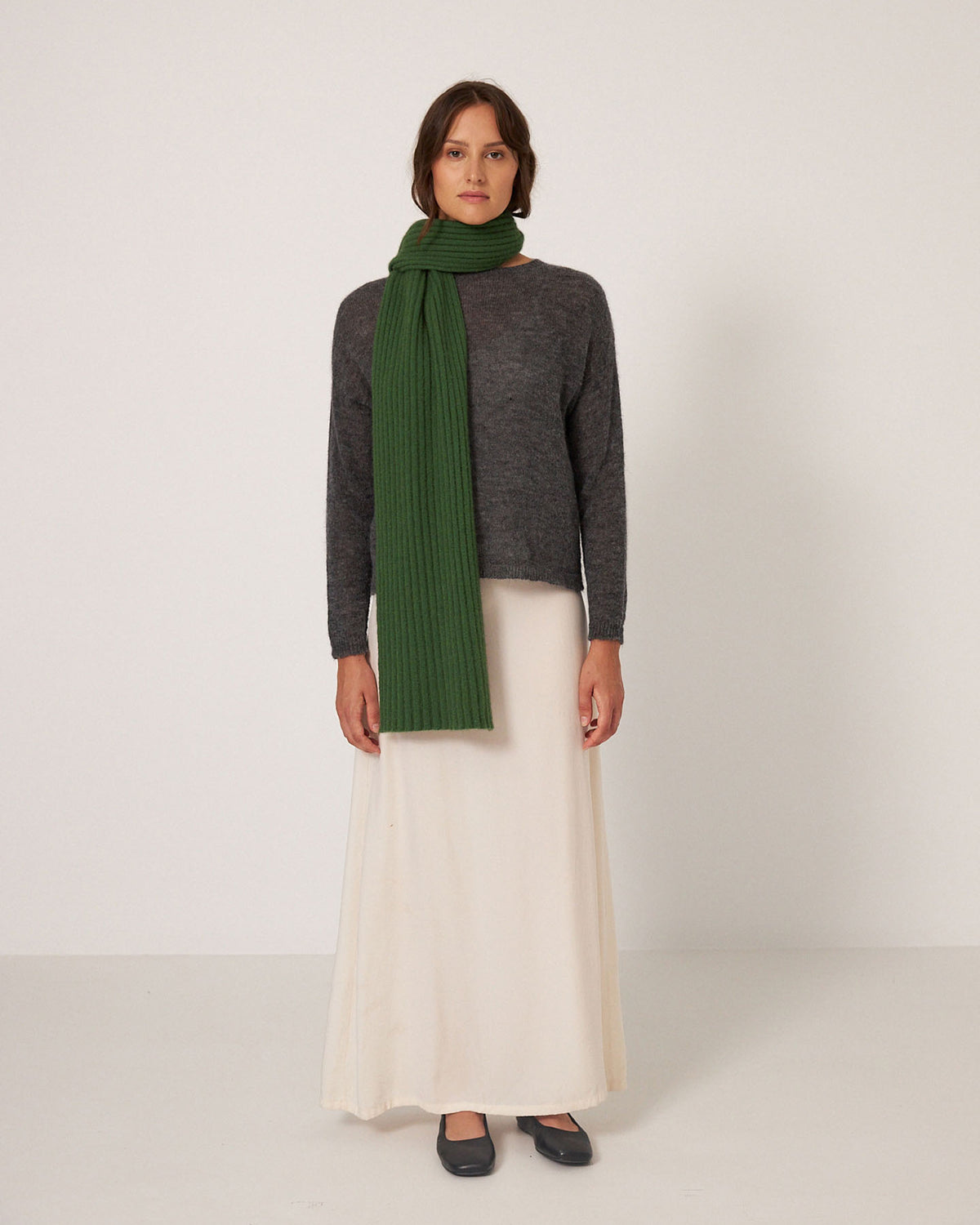 A person stands facing forward against a plain, light background, wearing a gray sweater, long cream skirt, black shoes, and the Francie Ripple Scarf – Herb Green (170cm at widest point).