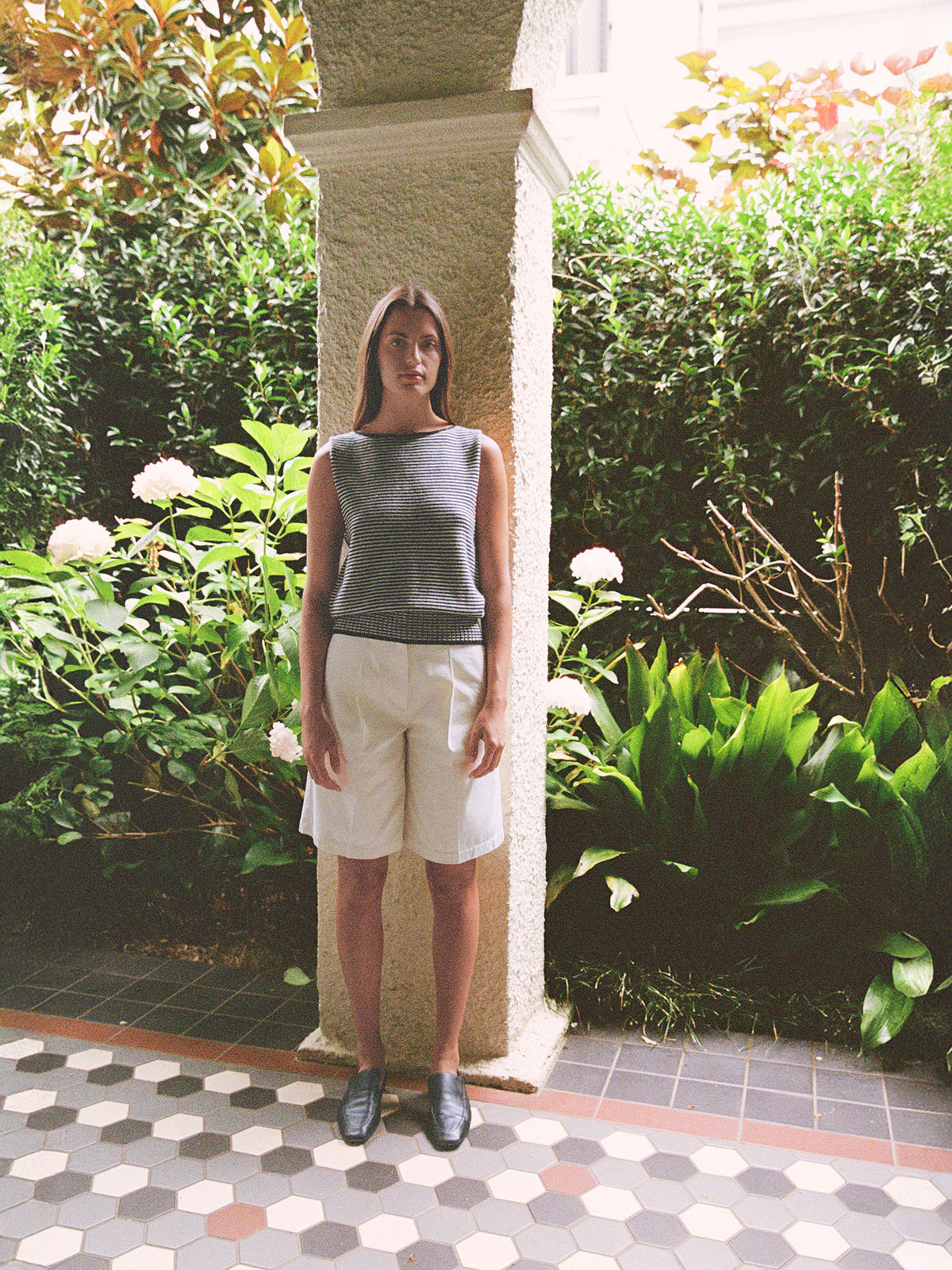 A woman stands by a column in a garden, wearing the Francie Striped Star Vest – Black &amp; Creme. She pairs the sleeveless top with white shorts and black shoes, surrounded by lush green plants and flowers.