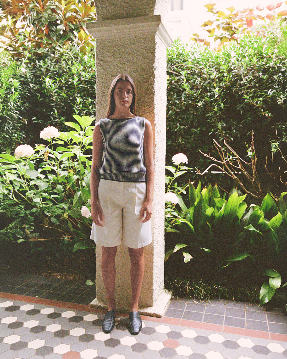 A woman stands by a column in a garden, wearing the Francie Striped Star Vest – Black & Creme. She pairs the sleeveless top with white shorts and black shoes, surrounded by lush green plants and flowers.