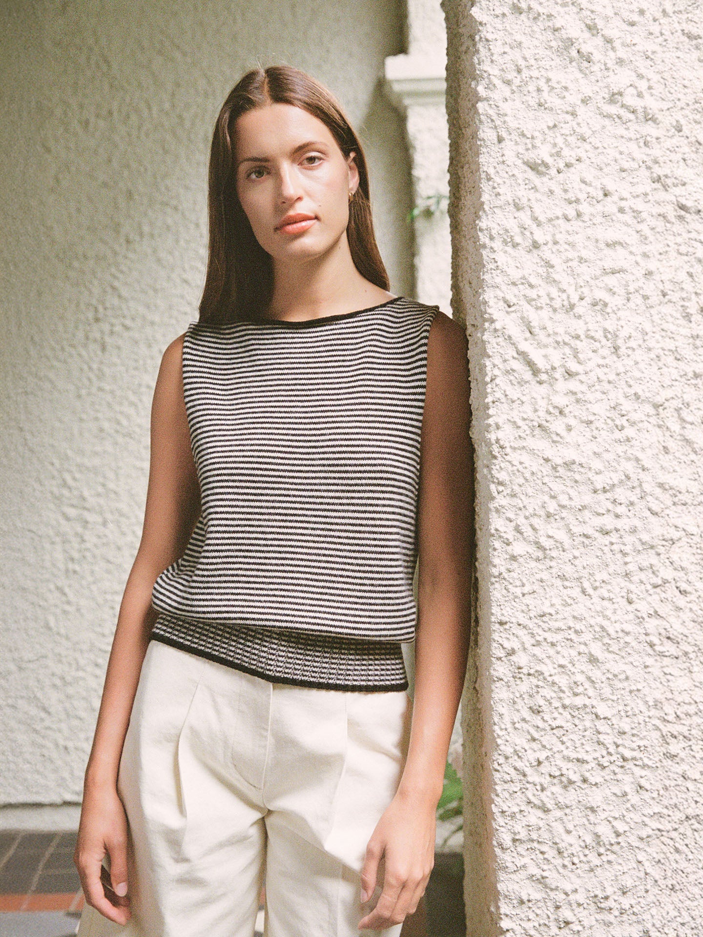 A woman with long brown hair leans against a textured wall, wearing Francie’s Striped Star Vest in black & creme and white pants.