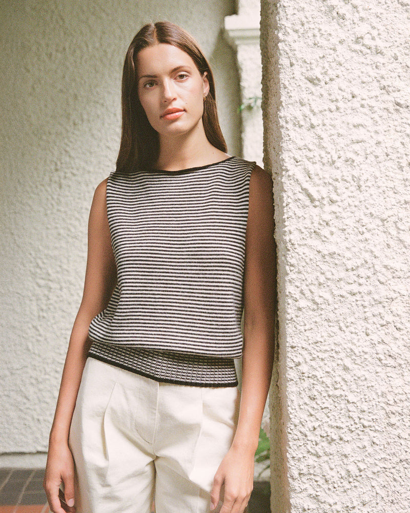 A woman with long brown hair leans against a textured wall, wearing Francie’s Striped Star Vest in black & creme and white pants.