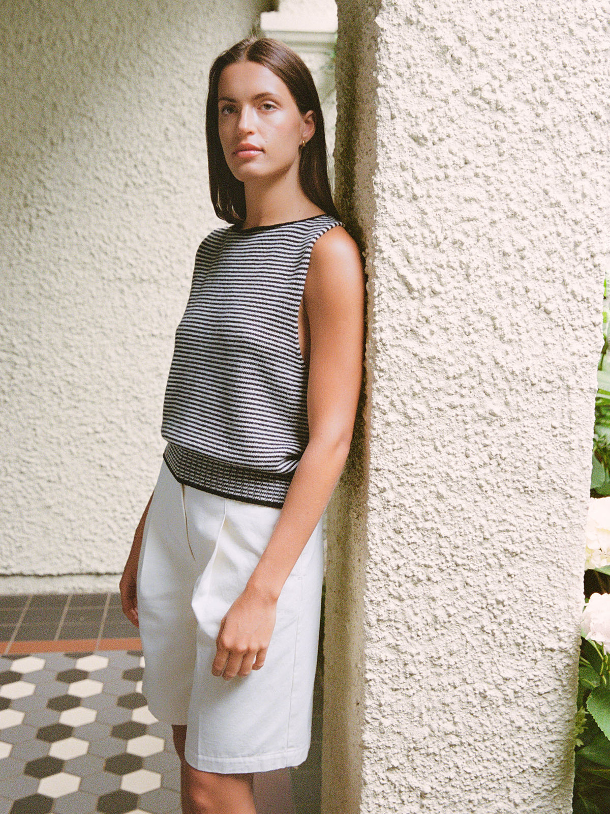 A woman wears the Francie Striped Star Vest – Black &amp; Creme with white shorts, standing by a textured white pillar with greenery in the background.