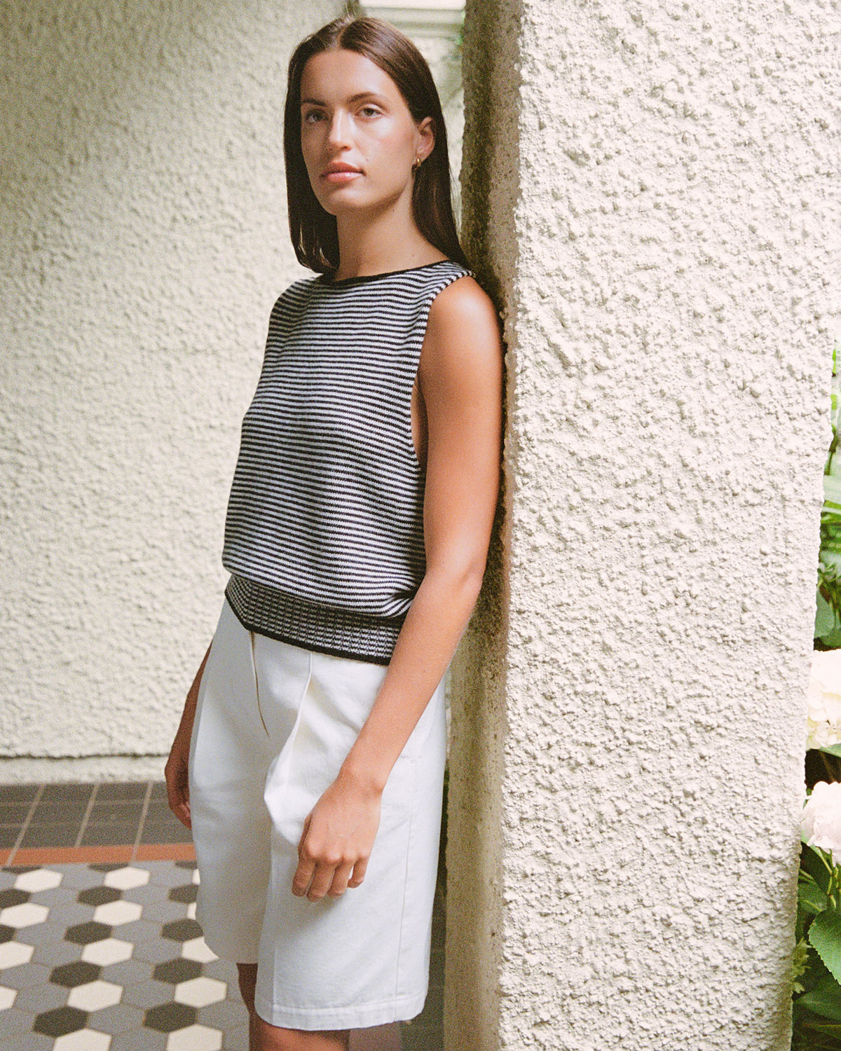 A woman wears the Francie Striped Star Vest – Black & Creme with white shorts, standing by a textured white pillar with greenery in the background.