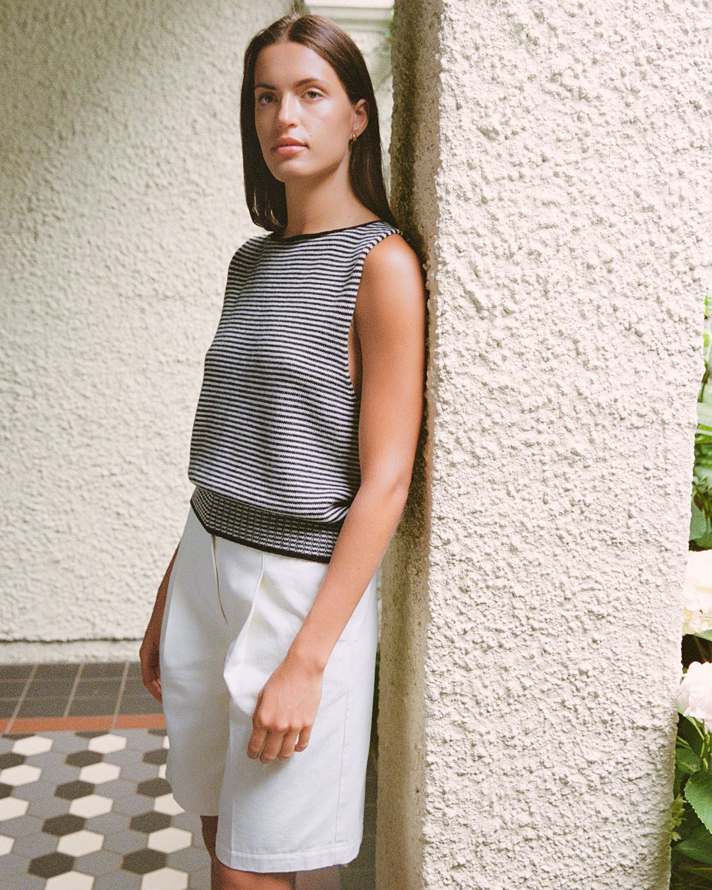 A woman with long brown hair leans against a textured wall, wearing Francie’s Striped Star Vest in black & creme and white pants.