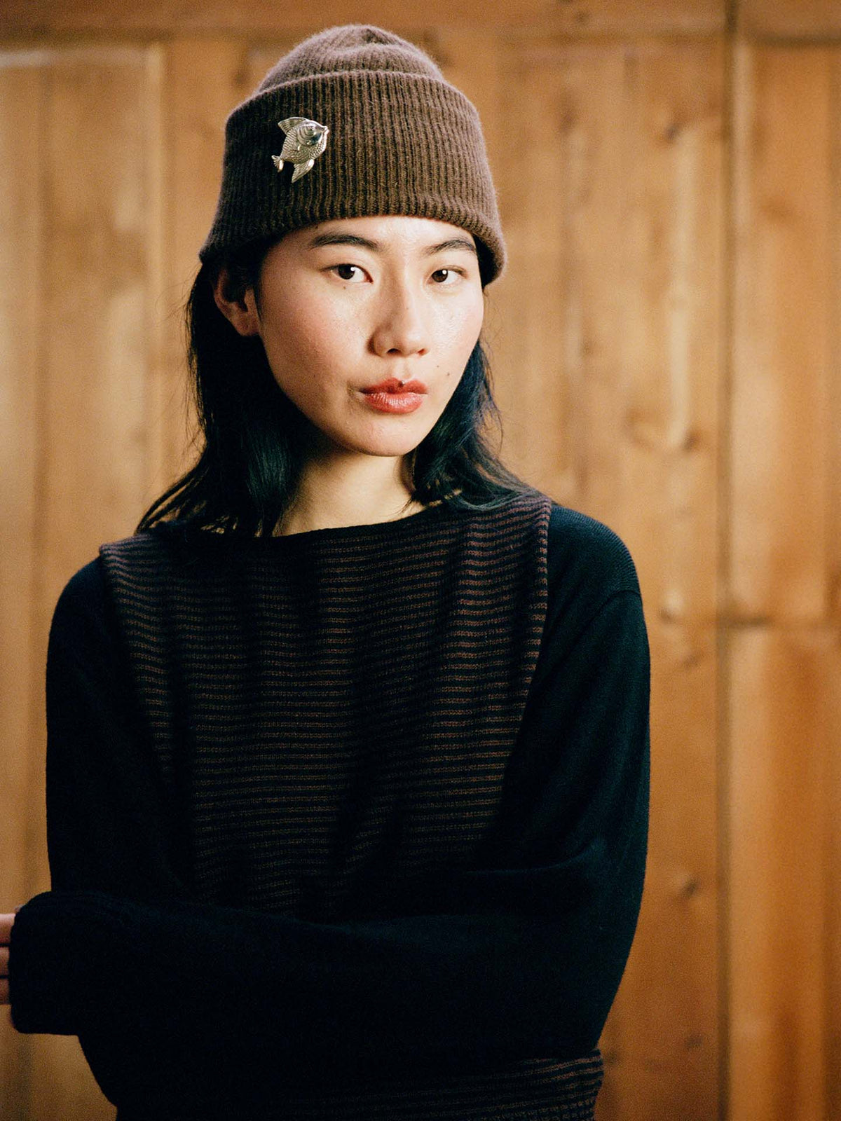 A woman stands in front of a wooden wall, looking at the camera with her arms crossed, wearing the Striped Star Vest in Chestnut & Black and a brown knit hat.