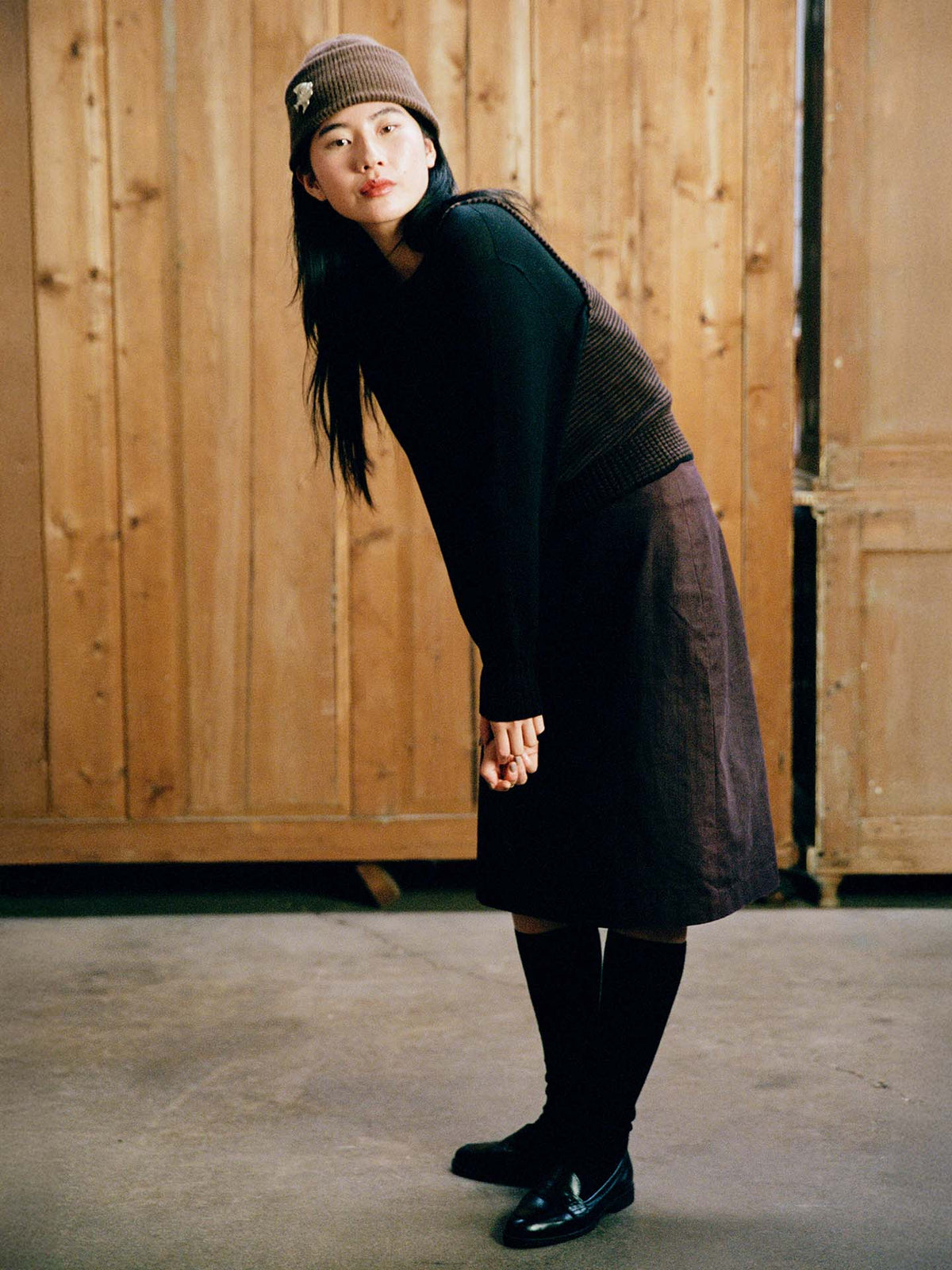 A woman poses indoors against a wooden background, wearing a relaxed fit brown skirt, black shoes, and the Striped Star Vest in Chestnut & Black. She bends forward slightly for the photo.