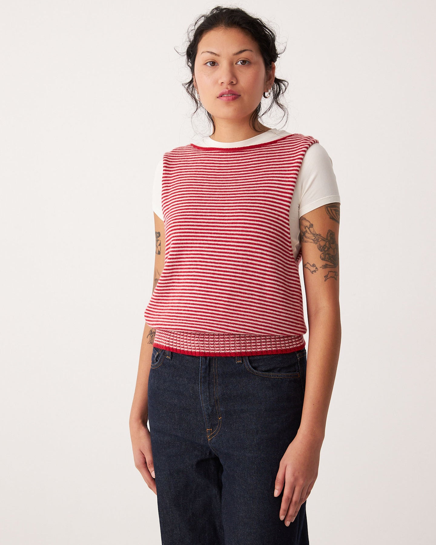 A woman stands against a plain background wearing Francie's Striped Star Vest – Cherry & Creme over a white short-sleeve shirt and dark jeans, creating a casual, petite-friendly look.