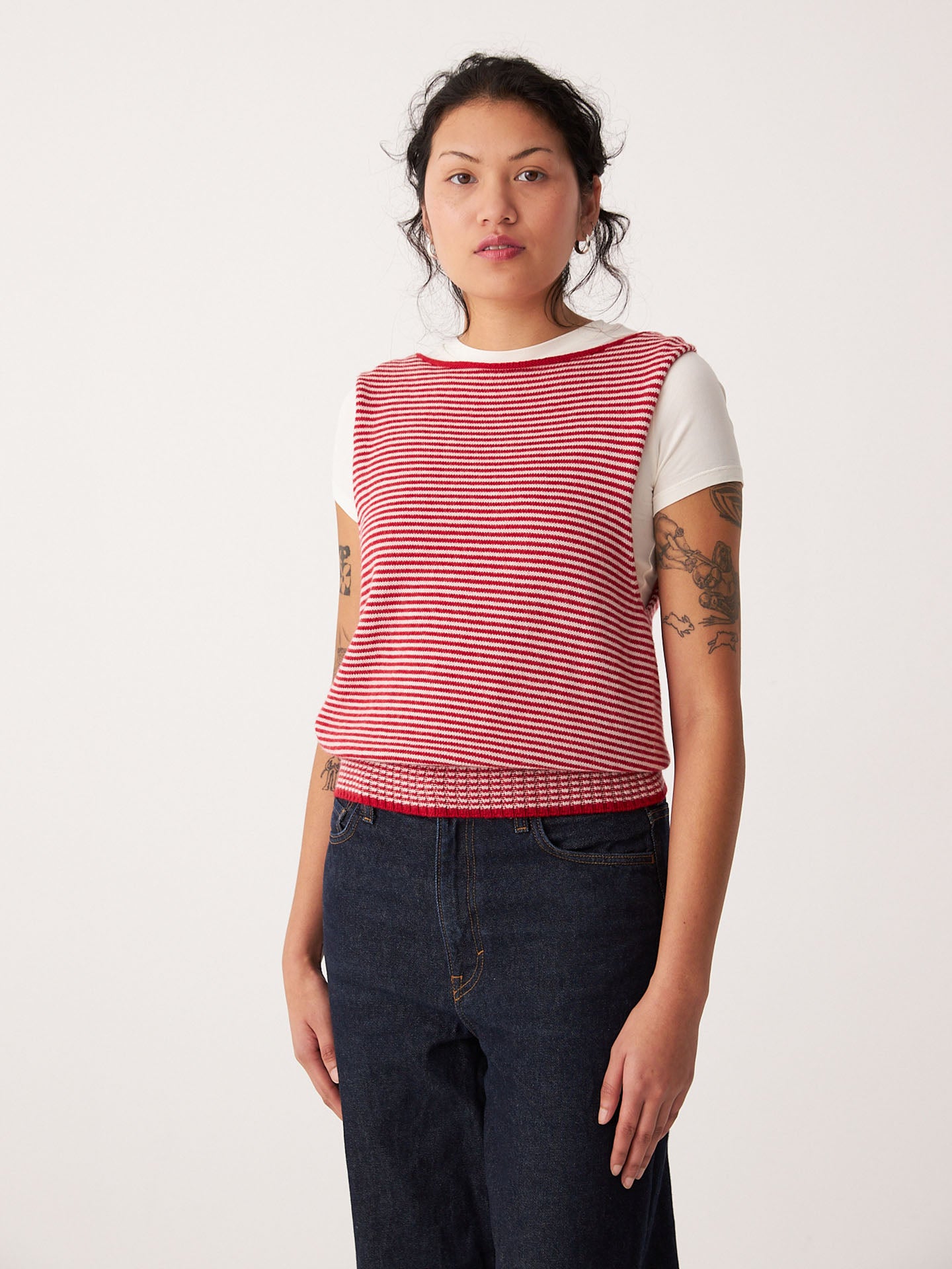 A woman stands against a plain background wearing Francie's Striped Star Vest – Cherry & Creme over a white short-sleeve shirt and dark jeans, creating a casual, petite-friendly look.