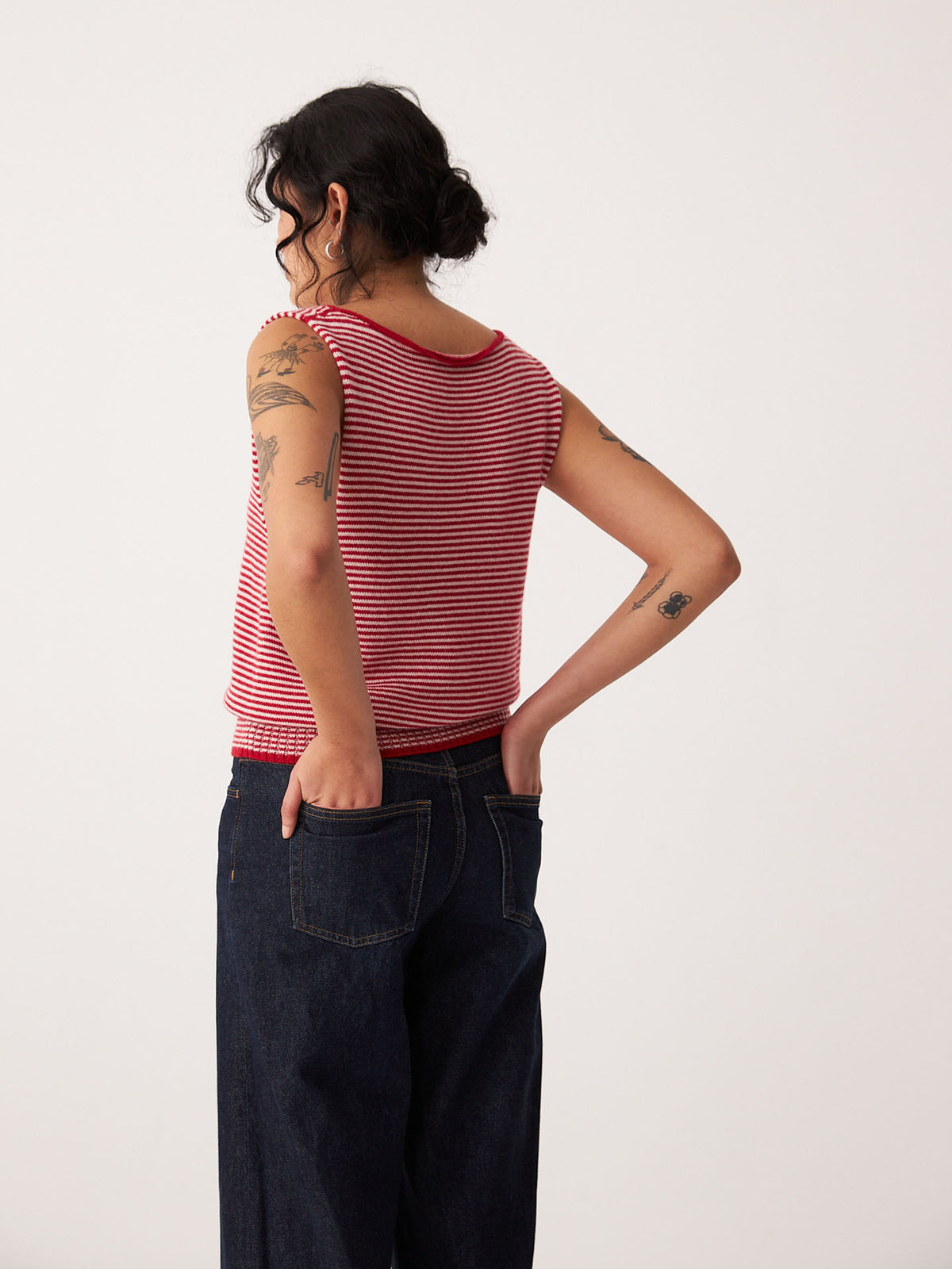 A petite person with tattoos wears the Francie Striped Star Vest – Cherry &amp; Creme with dark jeans, standing hands in back pockets, facing away from the camera.
