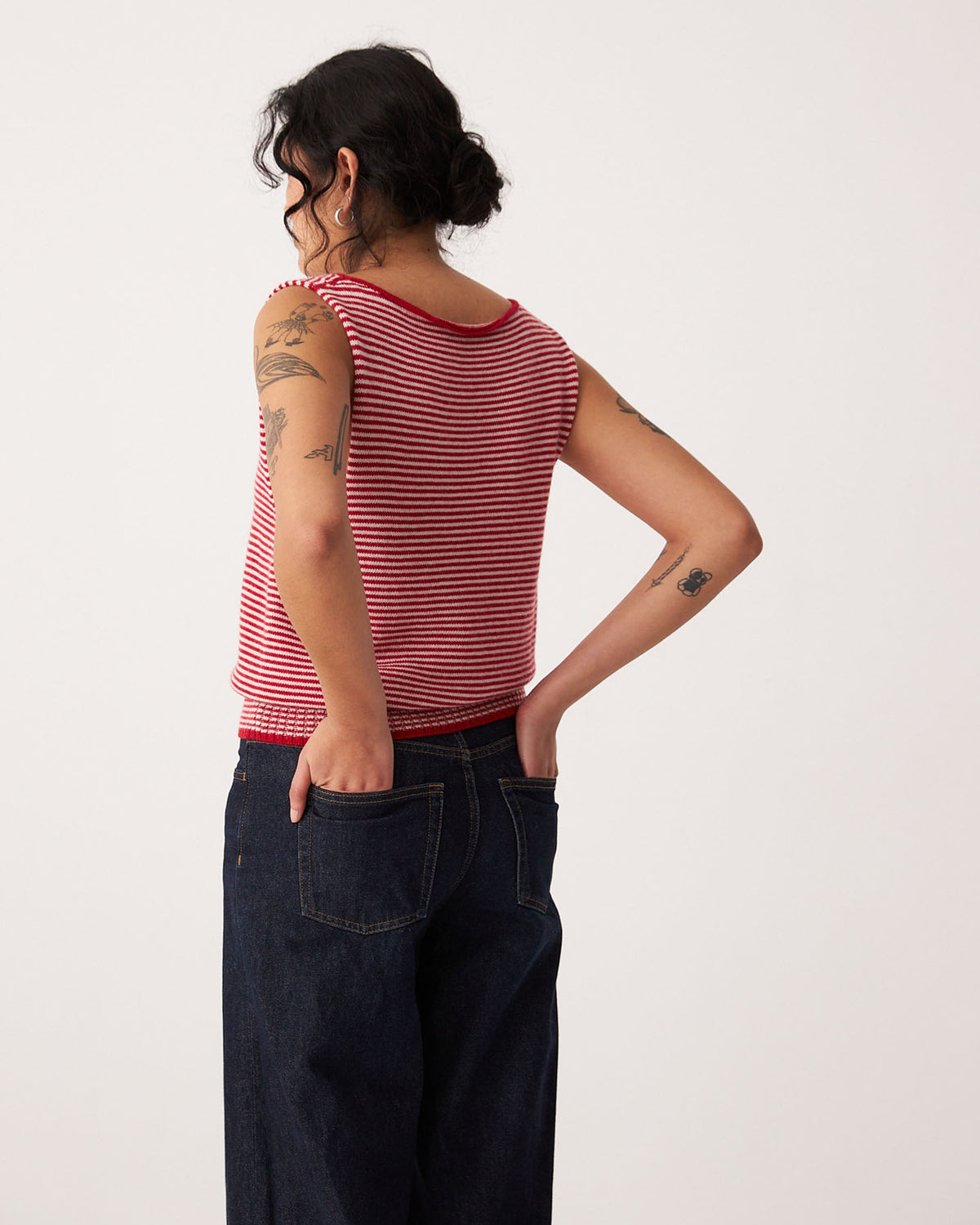 A petite person with tattoos wears the Francie Striped Star Vest – Cherry & Creme with dark jeans, standing hands in back pockets, facing away from the camera.