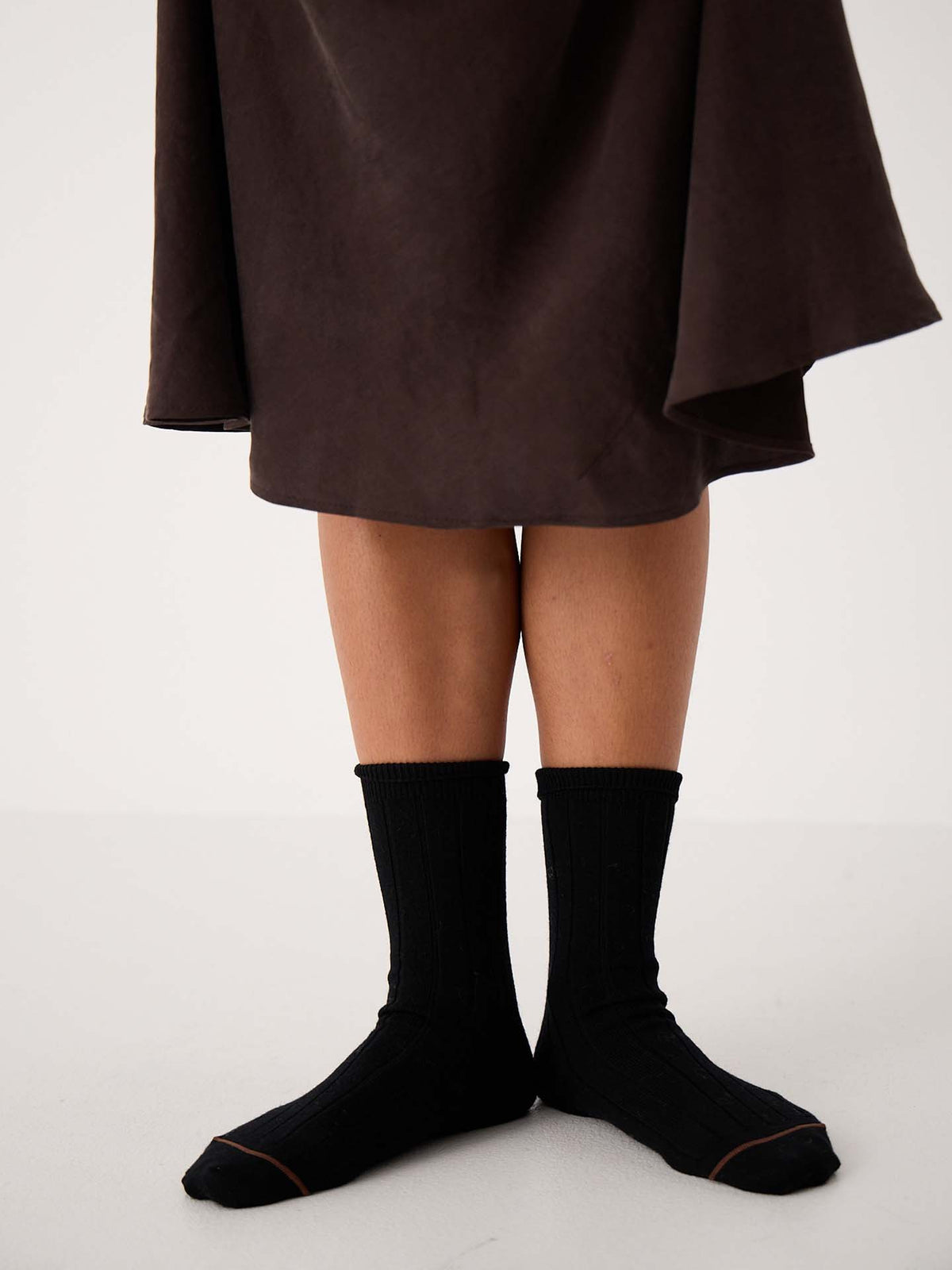 A person stands on a white floor wearing Tip Toe Crew Socks – Black & Chestnut Trim and a dark brown skirt; only legs and feet are visible, ideal for displaying various shoe size chart options (small, medium, large in EU, UK, US sizes).