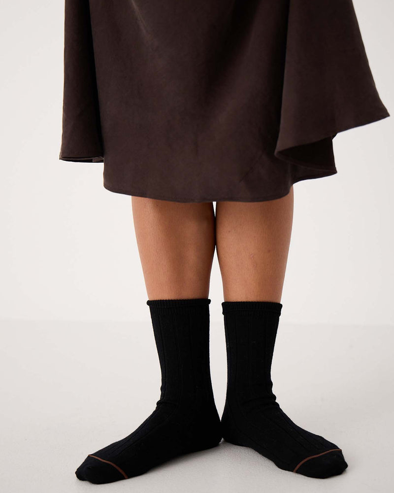 A person stands on a white floor wearing Tip Toe Crew Socks – Black & Chestnut Trim and a dark brown skirt; only legs and feet are visible, ideal for displaying various shoe size chart options (small, medium, large in EU, UK, US sizes).