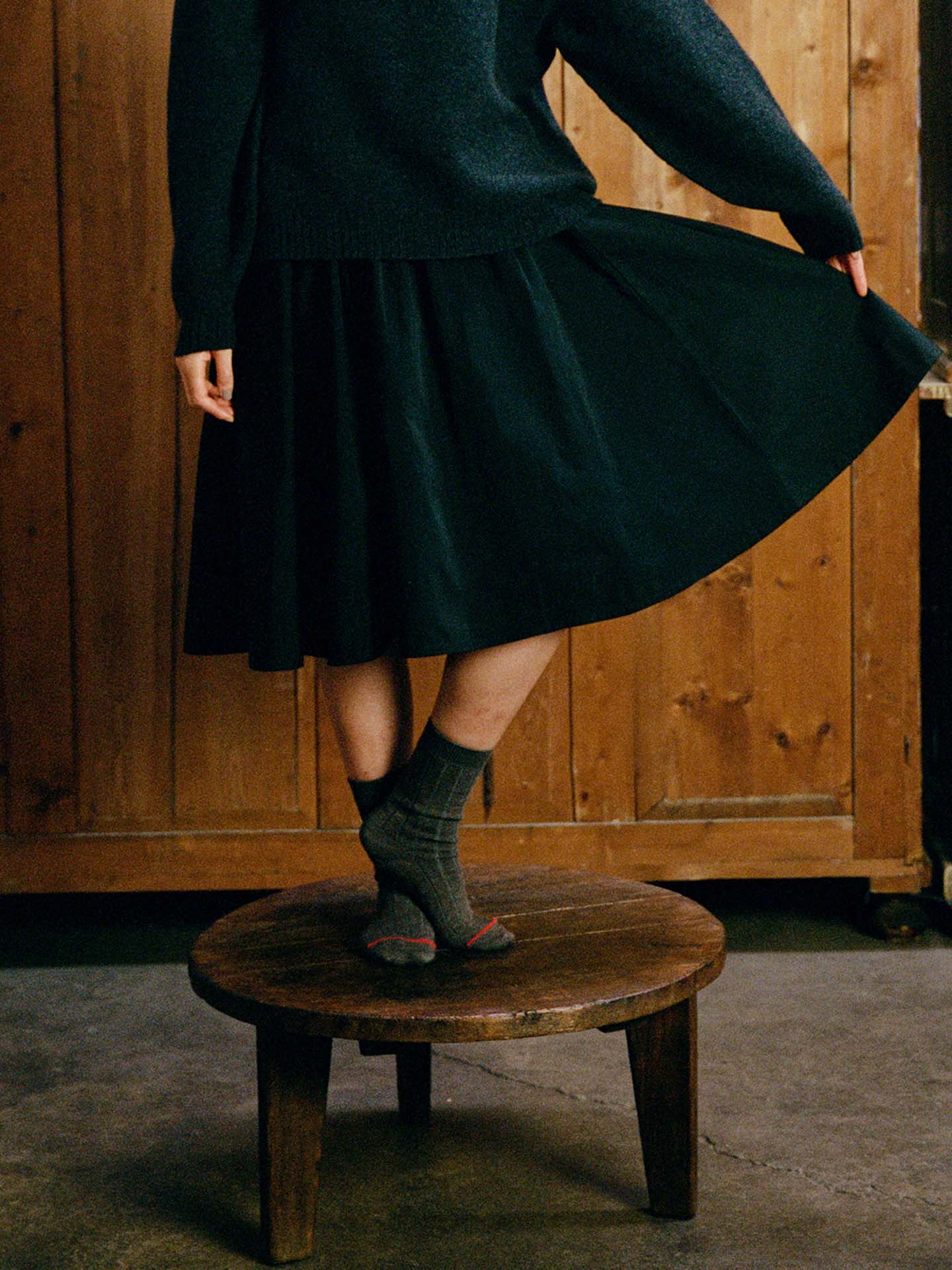 Person stands on a wooden stool, wearing a dark sweater, dark skirt, and Tip Toe Crew Socks – Grey & Cherry Trim, holding part of the skirt. Wooden wall in the background.