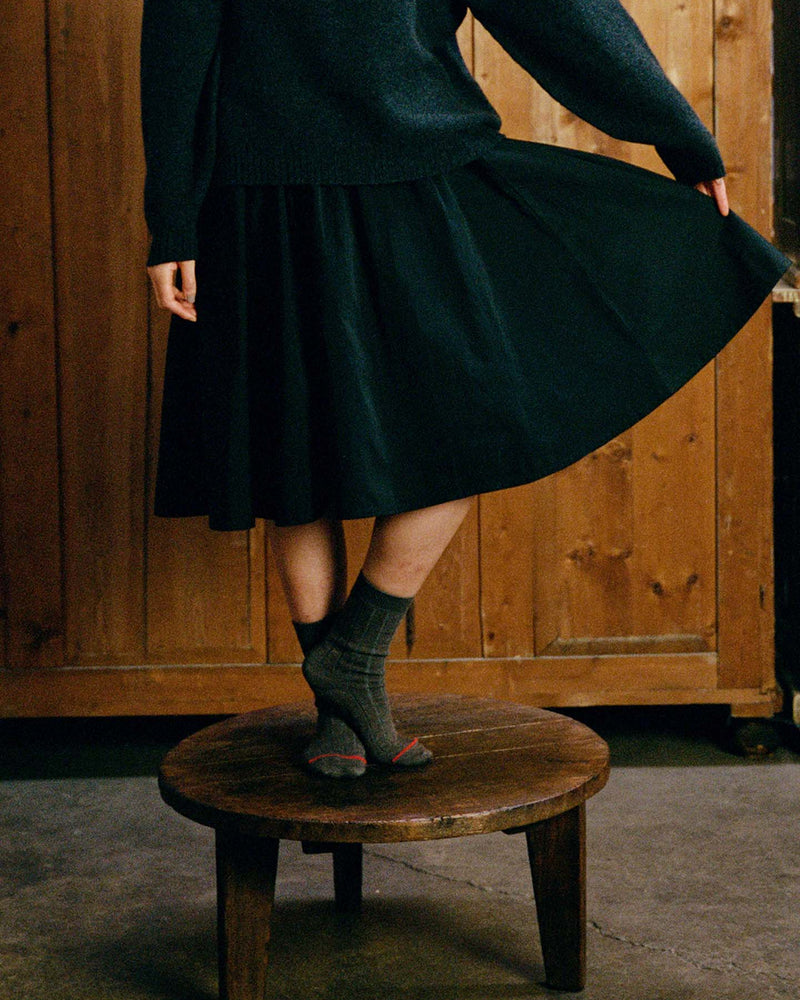Person stands on a wooden stool, wearing a dark sweater, dark skirt, and Tip Toe Crew Socks – Grey & Cherry Trim, holding part of the skirt. Wooden wall in the background.