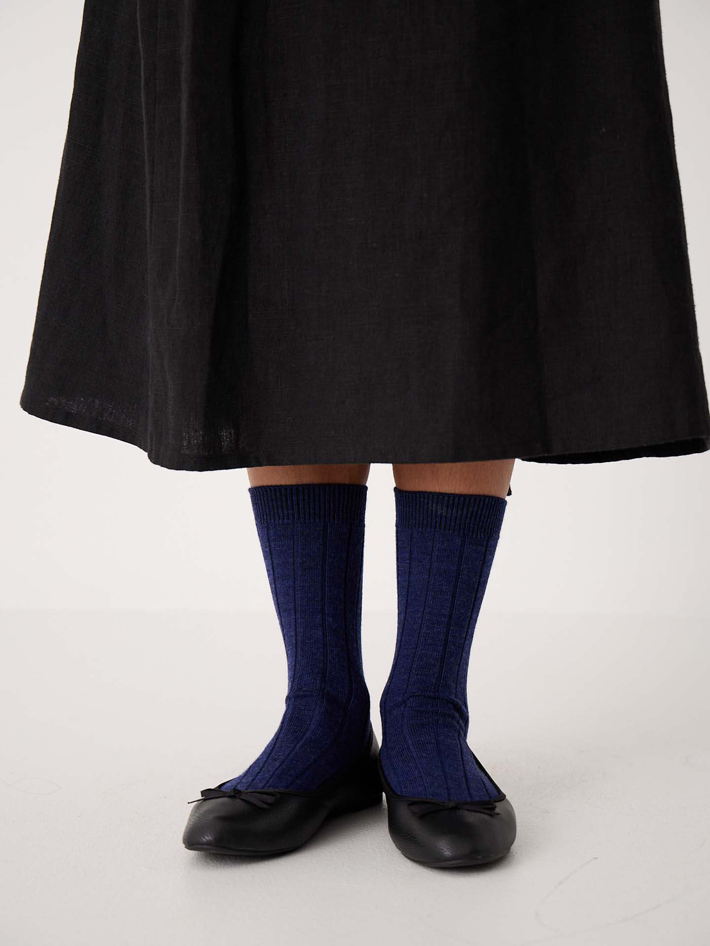 A person stands on a white floor wearing a long black skirt, black ballet flats, and Tip Toe Crew Socks in Sapphire with Creme Trim (available in sizes S, M, L) featuring a ribbed style.