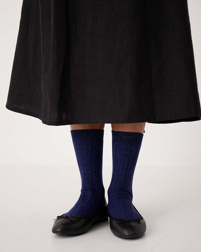 A person stands on a white floor wearing a long black skirt, black ballet flats, and Tip Toe Crew Socks in Sapphire with Creme Trim (available in sizes S, M, L) featuring a ribbed style.