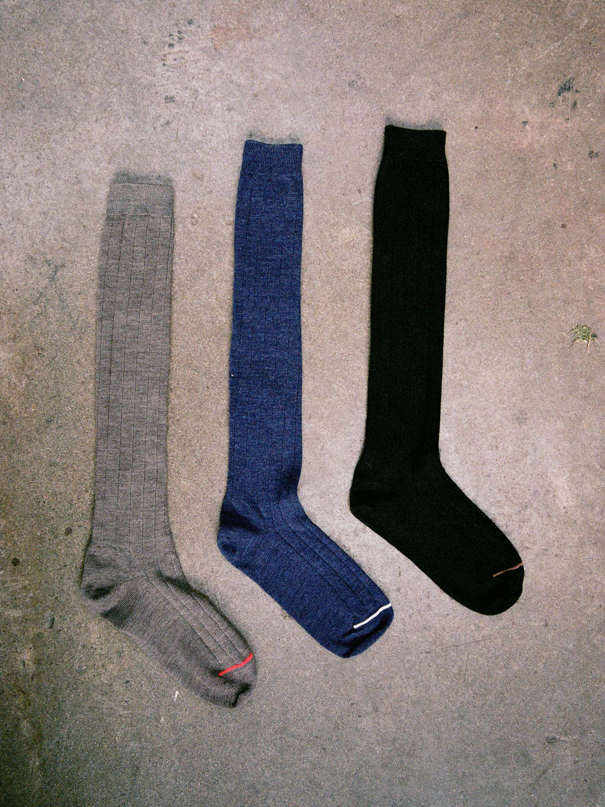 Three Tip Toe Knee High Socks in gray, blue, and black with chestnut trim are laid side by side on rough concrete. Check our size guide for your perfect fit using EU, UK, or US sizes.