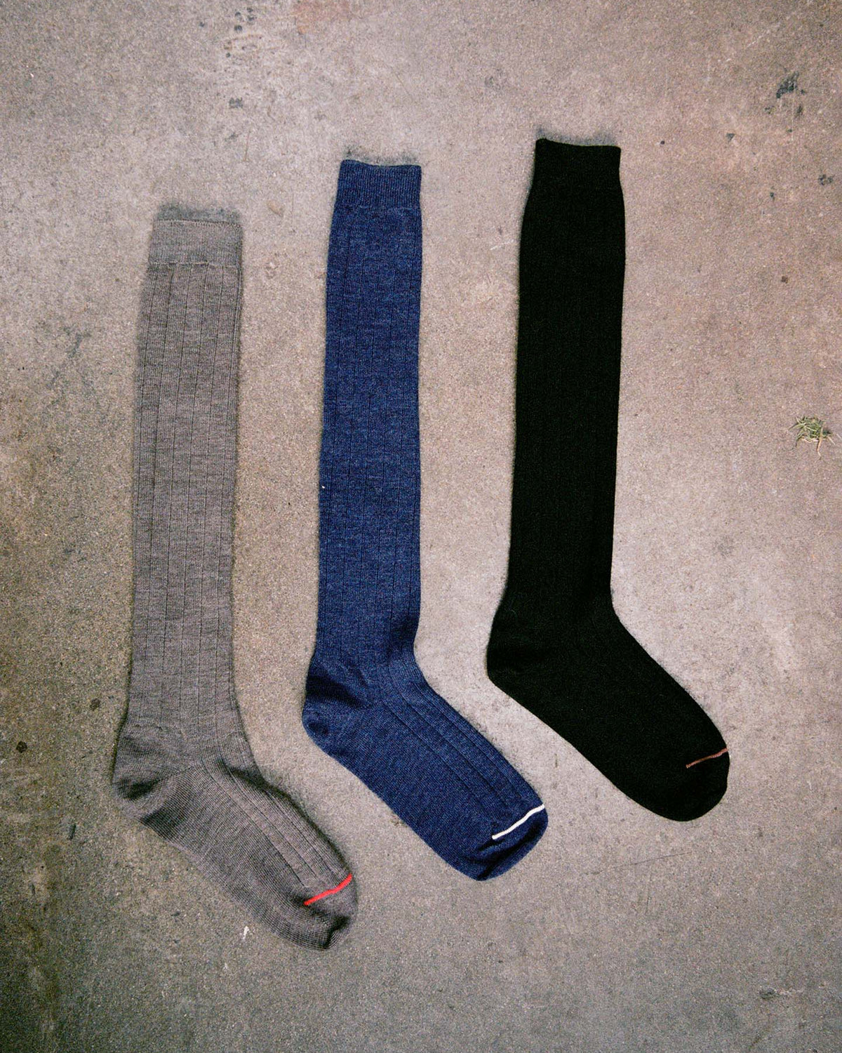 Three Tip Toe Knee High Socks in gray, blue, and black with chestnut trim are laid side by side on rough concrete. Check our size guide for your perfect fit using EU, UK, or US sizes.