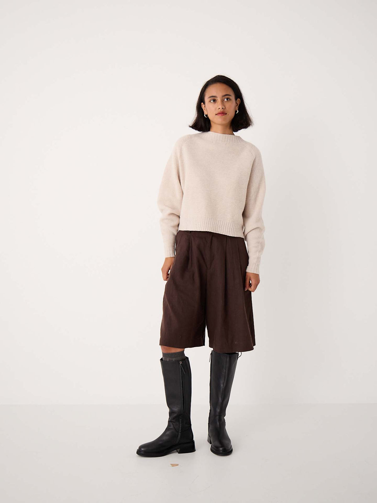 A woman stands against a plain white background wearing a cream sweater, brown knee-length shorts, black knee-high boots, and Tip Toe Knee High Socks in Grey & Cherry Trim.